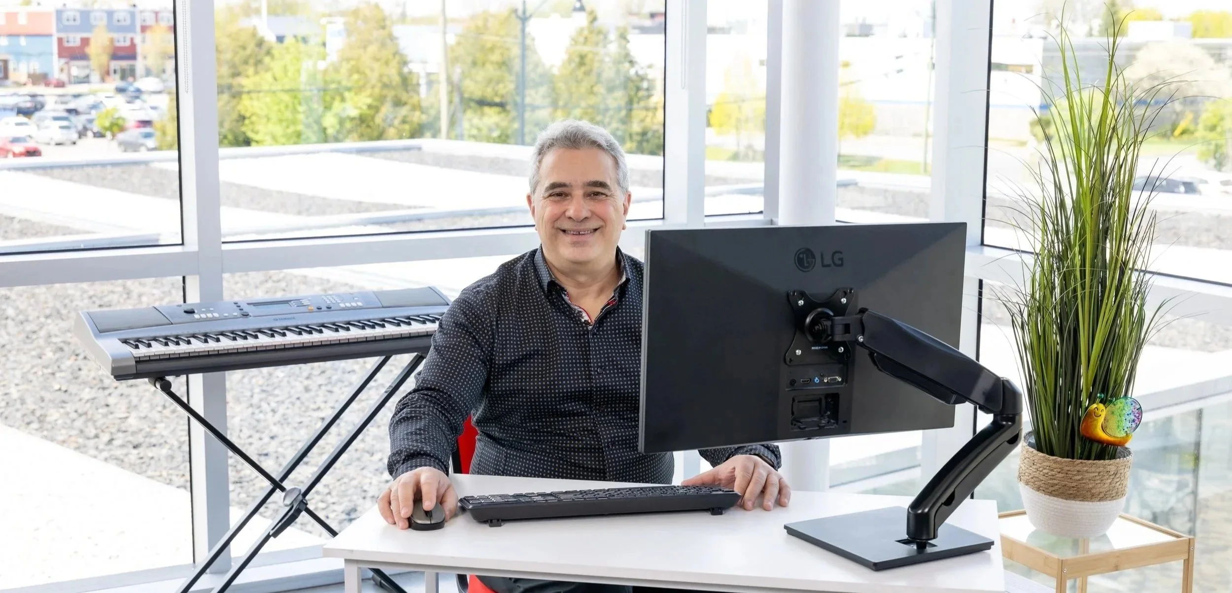 François Lessard, cofondateur et spécialiste, souriant, assis devant un ordinateur dans un bureau lumineux avec une grande fenêtre, piano électrique sur le côté, plante ornementale à côté, vue extérieure d'un parking et d'un quartier résidentiel.