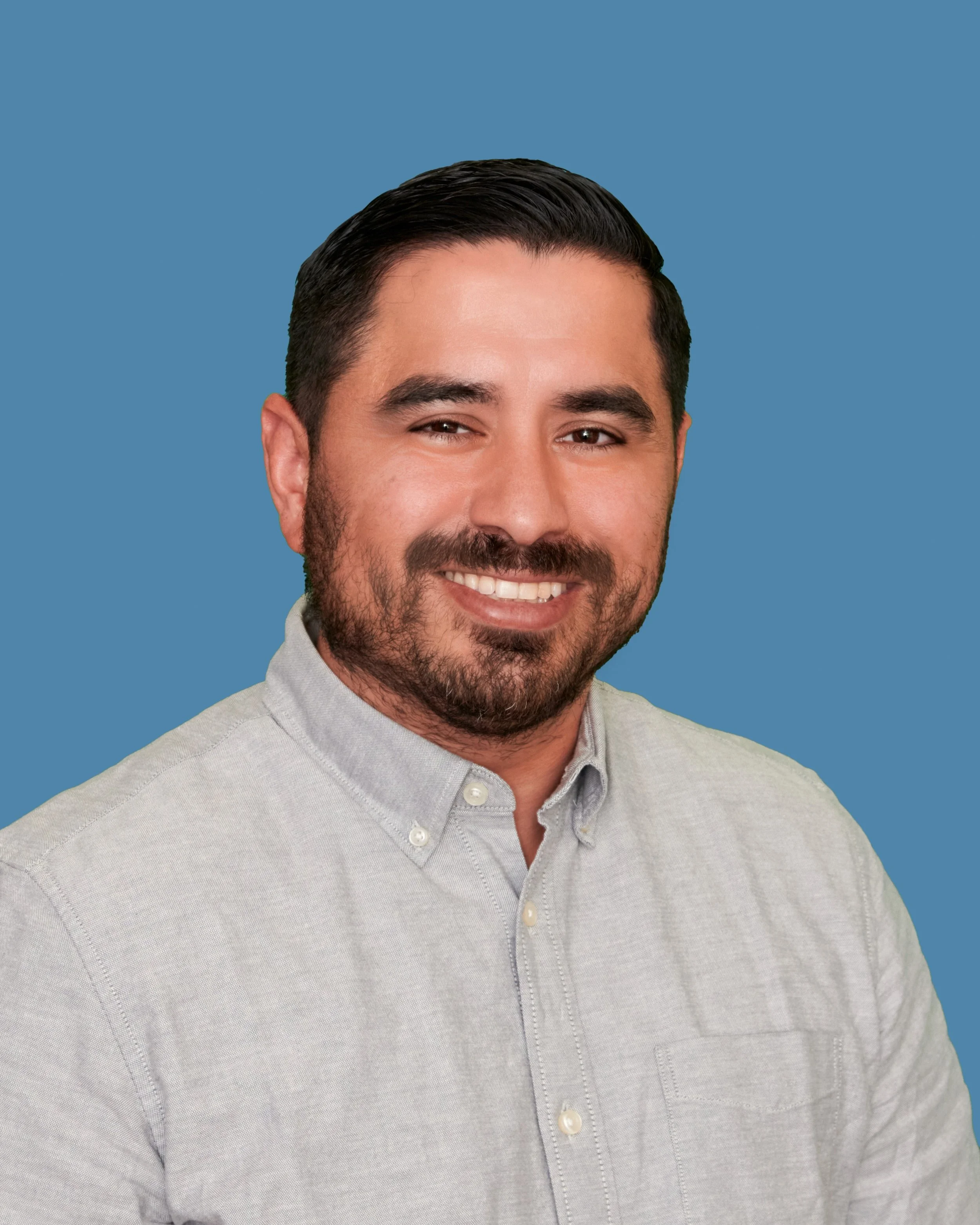 Headshot of a smiling man with dark hair and a beard, wearing a light gray button-up shirt against a solid blue background.