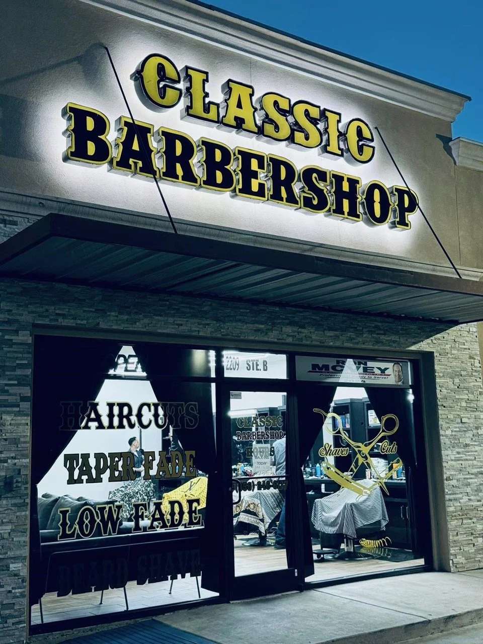 Exterior of a Classic Barbershop with a bright yellow and black sign reading 'Classic Barbershop'; storefront windows with promotional text and barber tools graphics, including scissors and a barber pole.