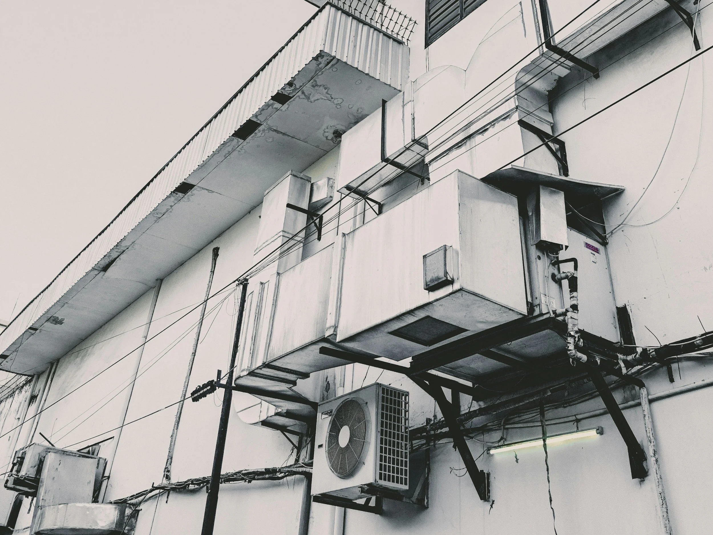 Close-up of a building's exterior showing various air conditioning units, vents, and electrical boxes attached to the white wall, with some visible wires and pipes.