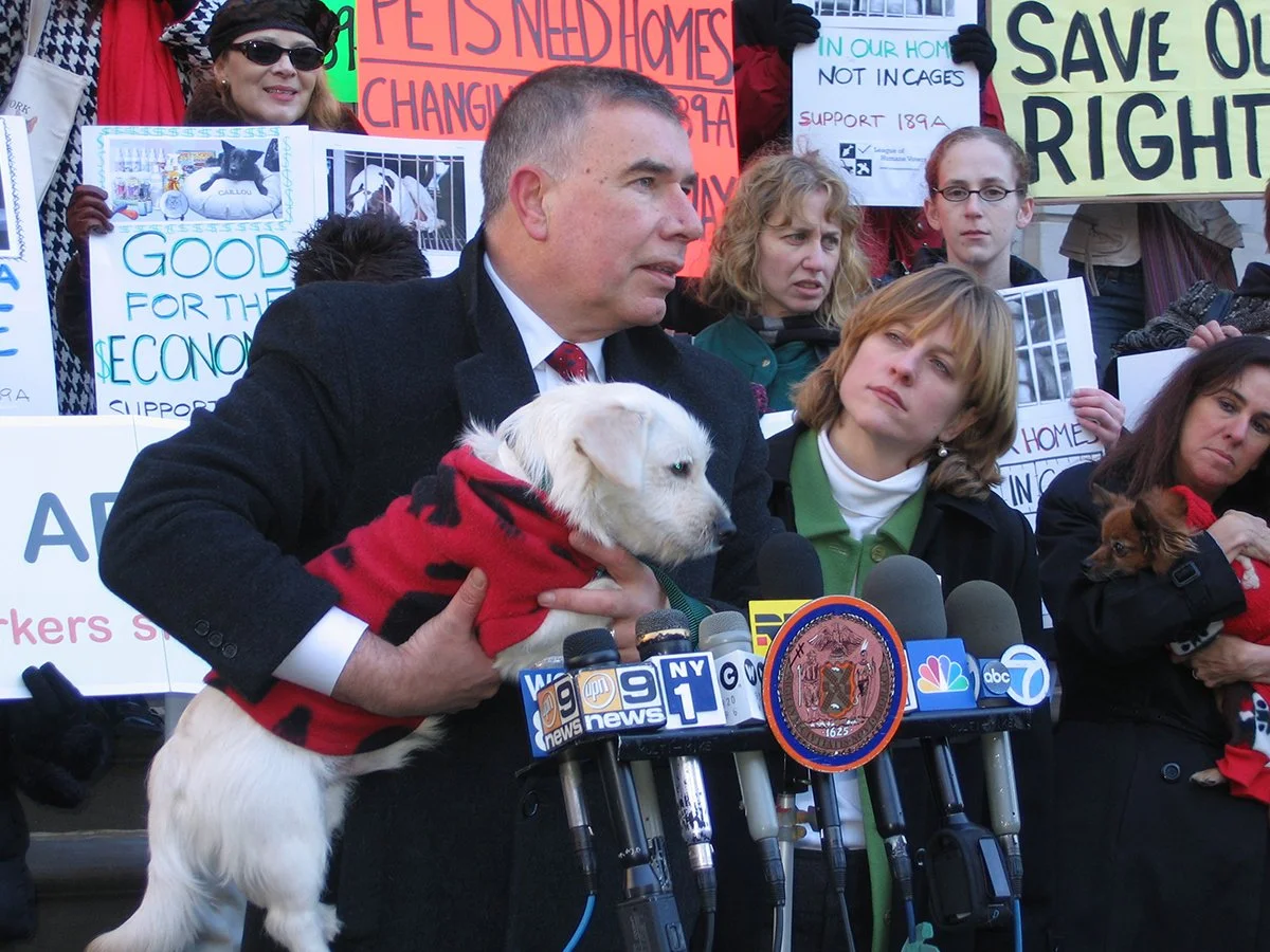 A group of people protesting with signs, some of which support pets' rights and oppose in cages. A man in a dark coat is speaking at a press conference, holding a white puppy wearing a red sweater with black spots. Multiple microphones are set up in front of him, and several women and children hold additional signs behind him.