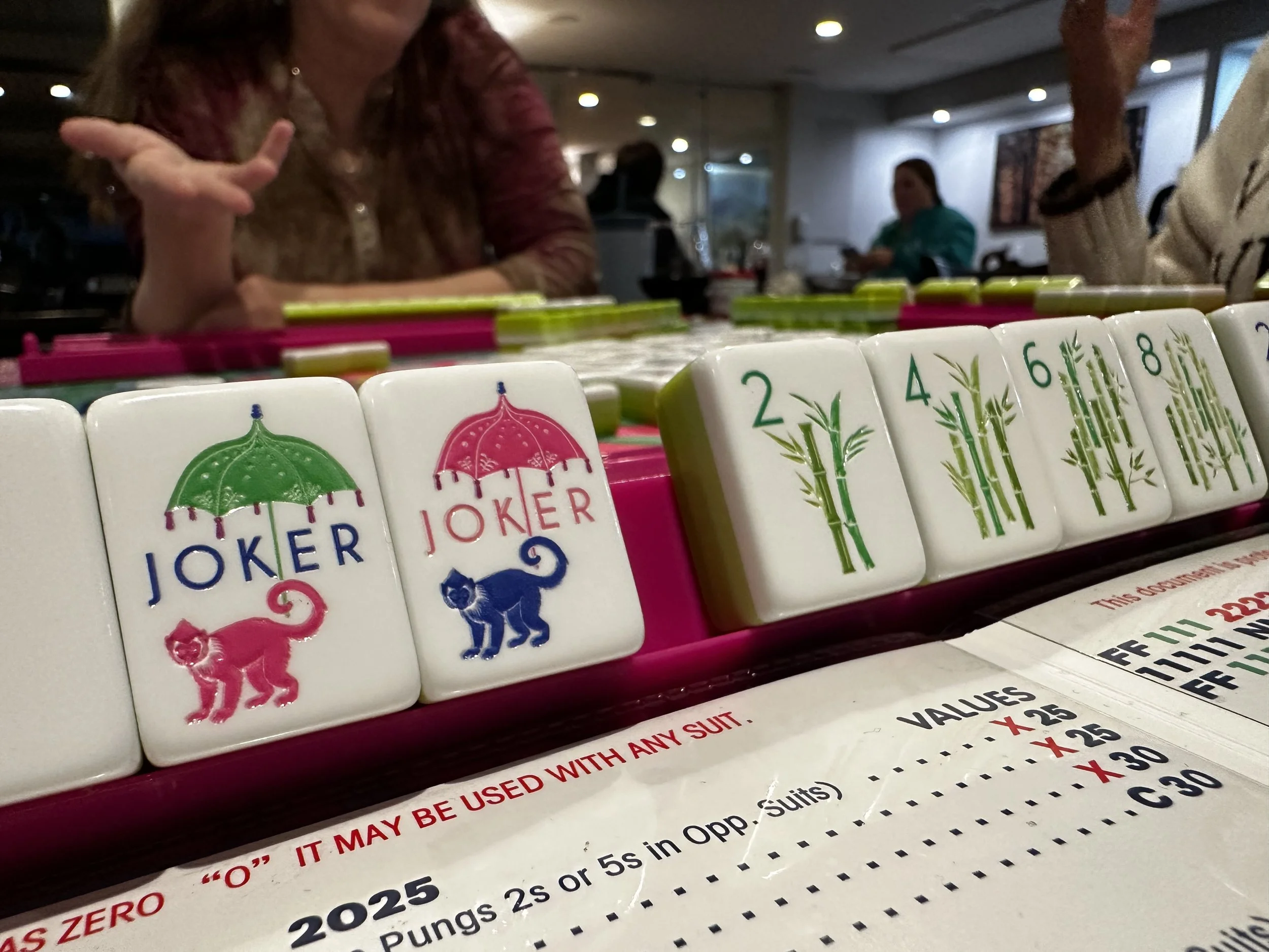 Close-up view of mahjong tiles, including two joker tiles with umbrella and cat symbols, and numbered bamboo tiles from 2 to 8, on a table with people in the background in a restaurant or café.