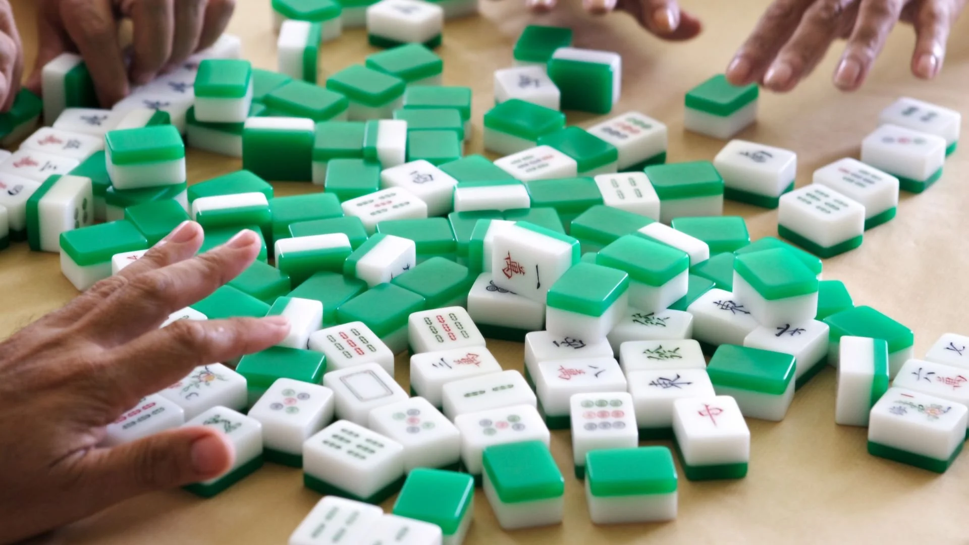 Multiple hands playing a game of Mahjong on a table with scattered green and white tiles.