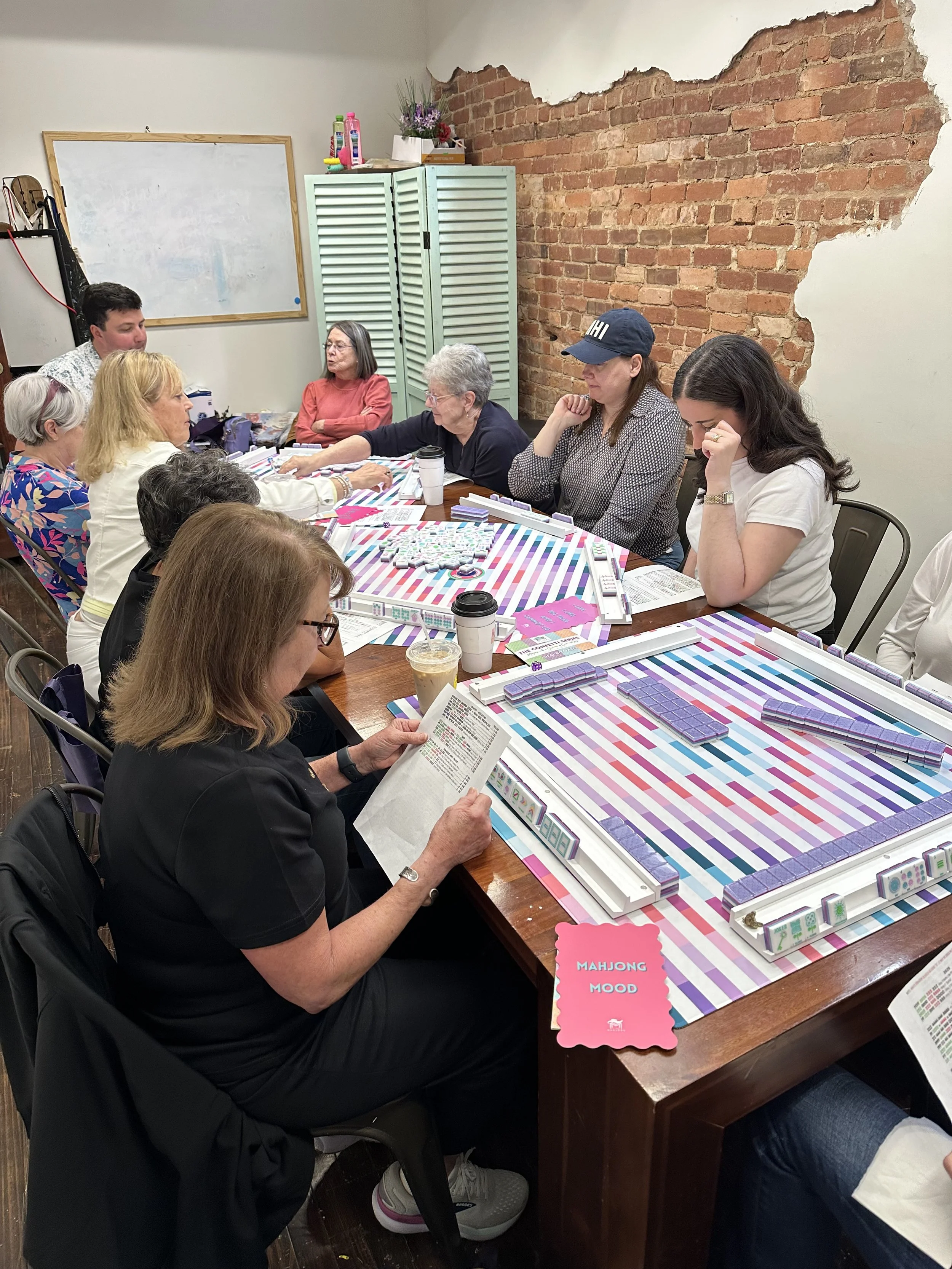A group of women and one man gathered around a table playing and learning Mahjong in a cozy room with exposed brick wall and colorful striped table surface.