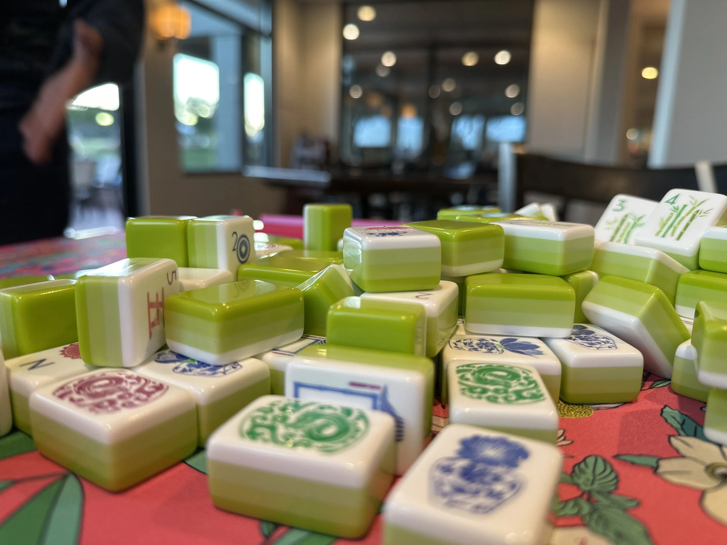 Close-up of Mahjong tiles spilling onto a colorful tablecloth, with a blurred background of a room and windows.