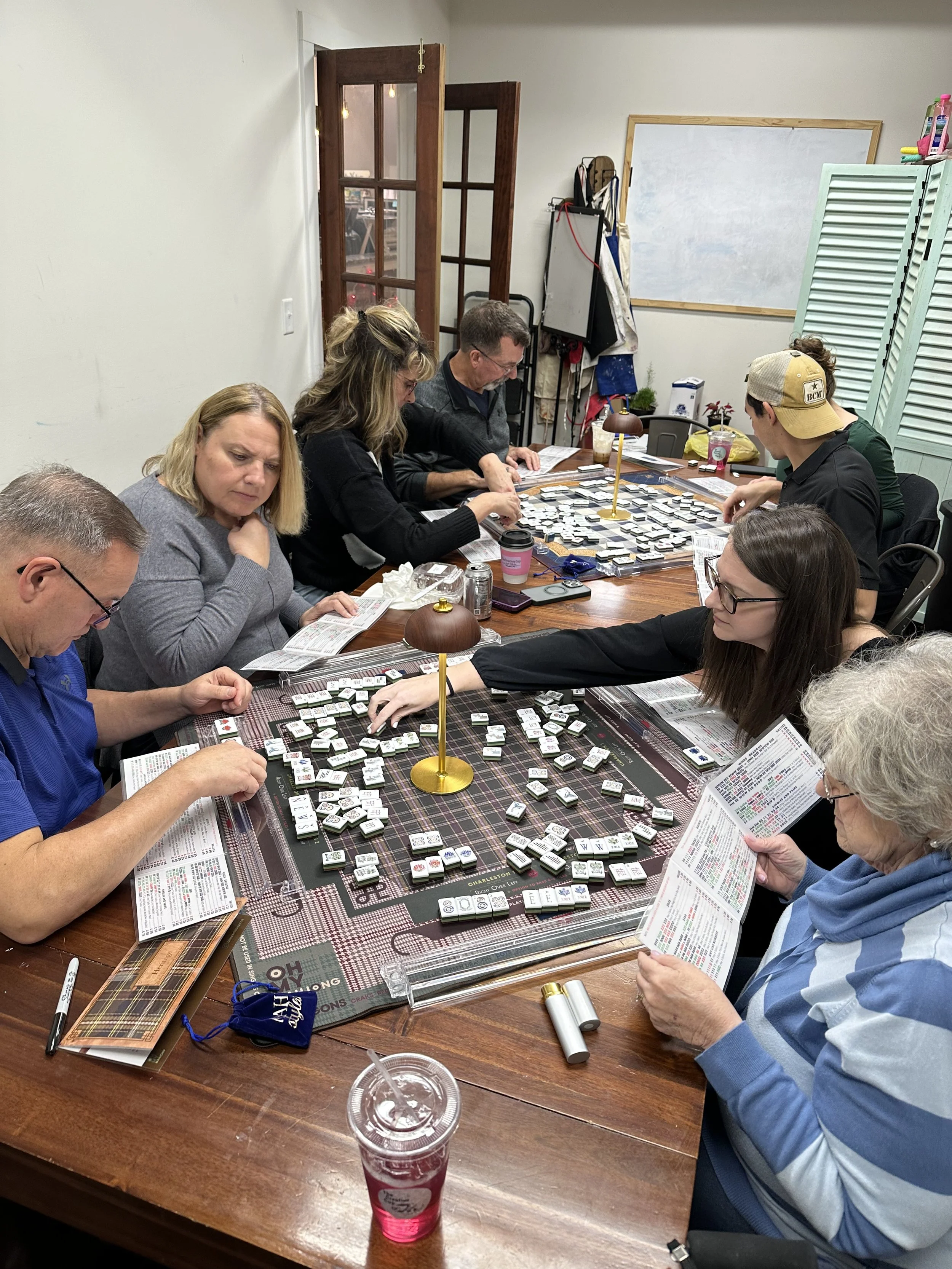 People playing a game of Mahjong around a wooden table, reading instruction booklets, with Mahjong tiles and accessories on the table.