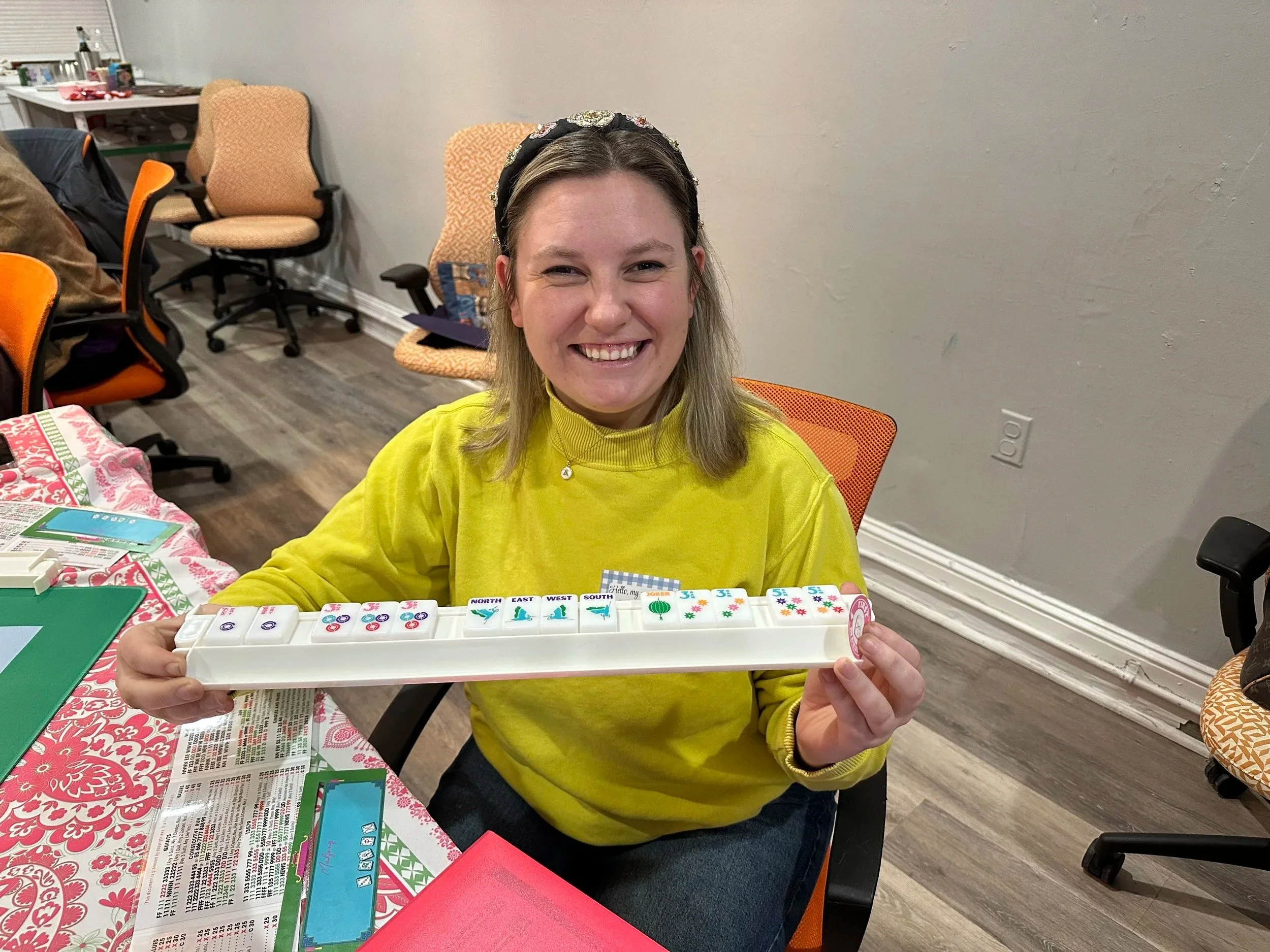 A woman wearing a yellow sweater and a floral headband is smiling and holding a white tray with colorful dice showing various numbers and symbols. She is sitting at a table with a pink and white tablecloth, in a room with multiple chairs and a gray wall.