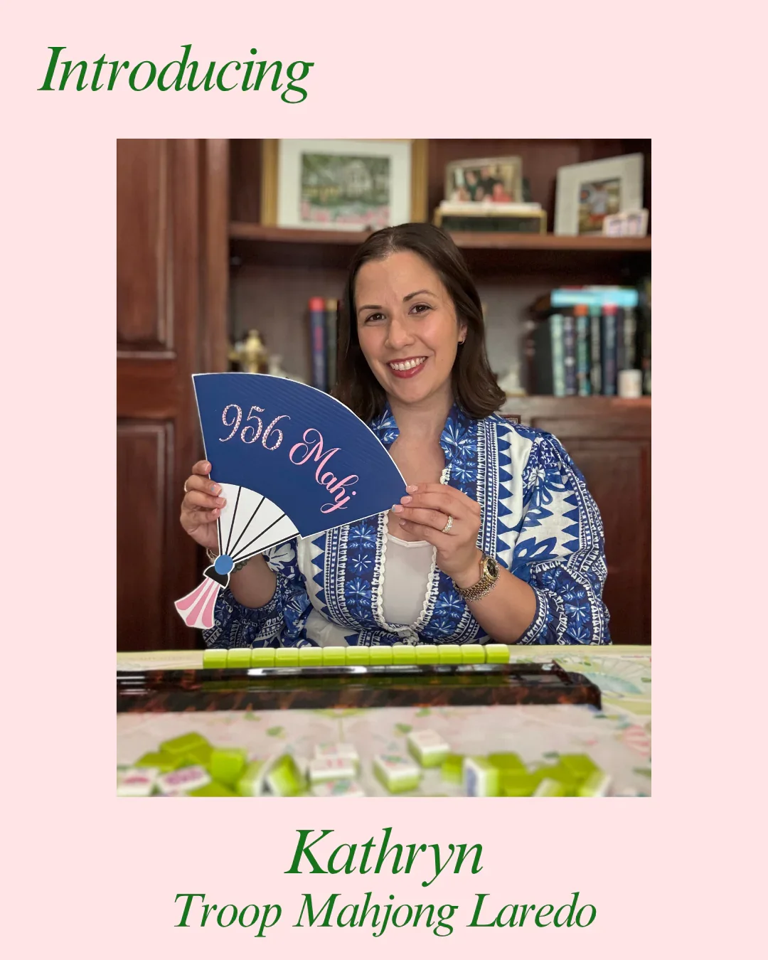 A woman named Kathryn holding a blue fan with '956 Mahaj' written on it, sitting at a table with green tiles and a board game, in a room with wooden shelves and framed pictures.