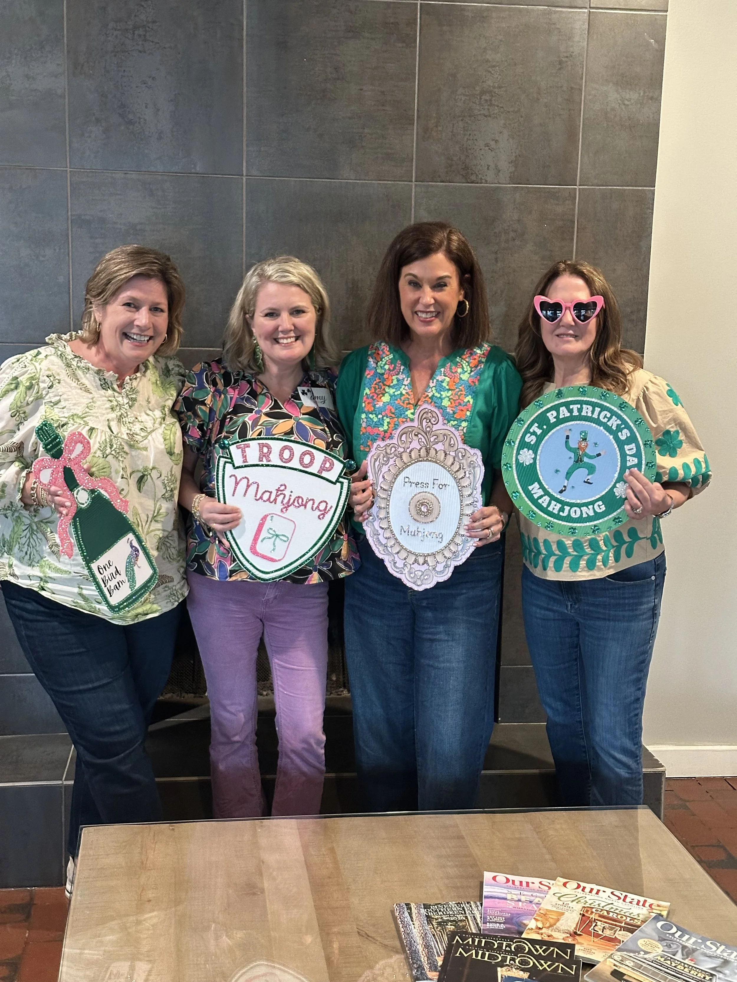 Four women standing together holding St. Patrick's Day-themed signs and decorations, smiling for the camera indoors.