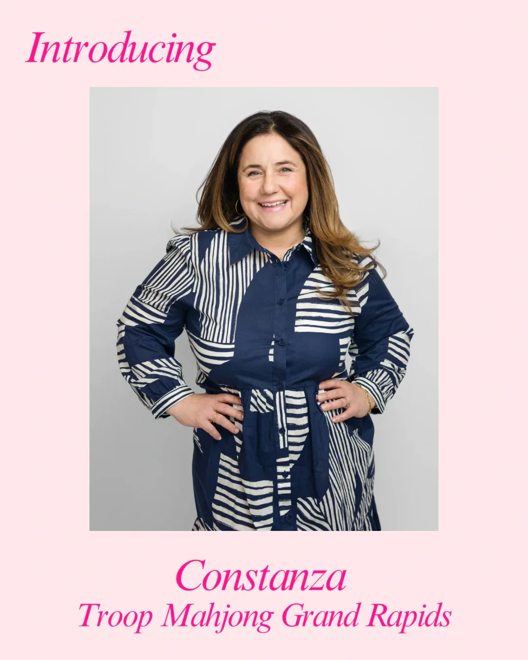 A woman with long wavy brown hair, wearing a navy and white patterned blouse, smiling with her hands on her hips, standing against a plain gray background. The image is part of a promotional graphic with pink text stating "Introducing" at the top and "Constanza Troop Mahjong Grand Rapids" at the bottom.