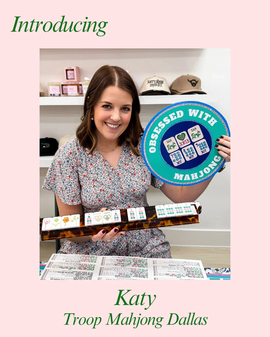 A woman smiling and holding a large round sign that says "Obsessed with Mahjong". She is sitting in front of a table with mahjong tiles and a sheet of paper. The background has shelves with hats and candles. The image has a pink background with green text that says "Introducing Katy Troop Mahjong Dallas".