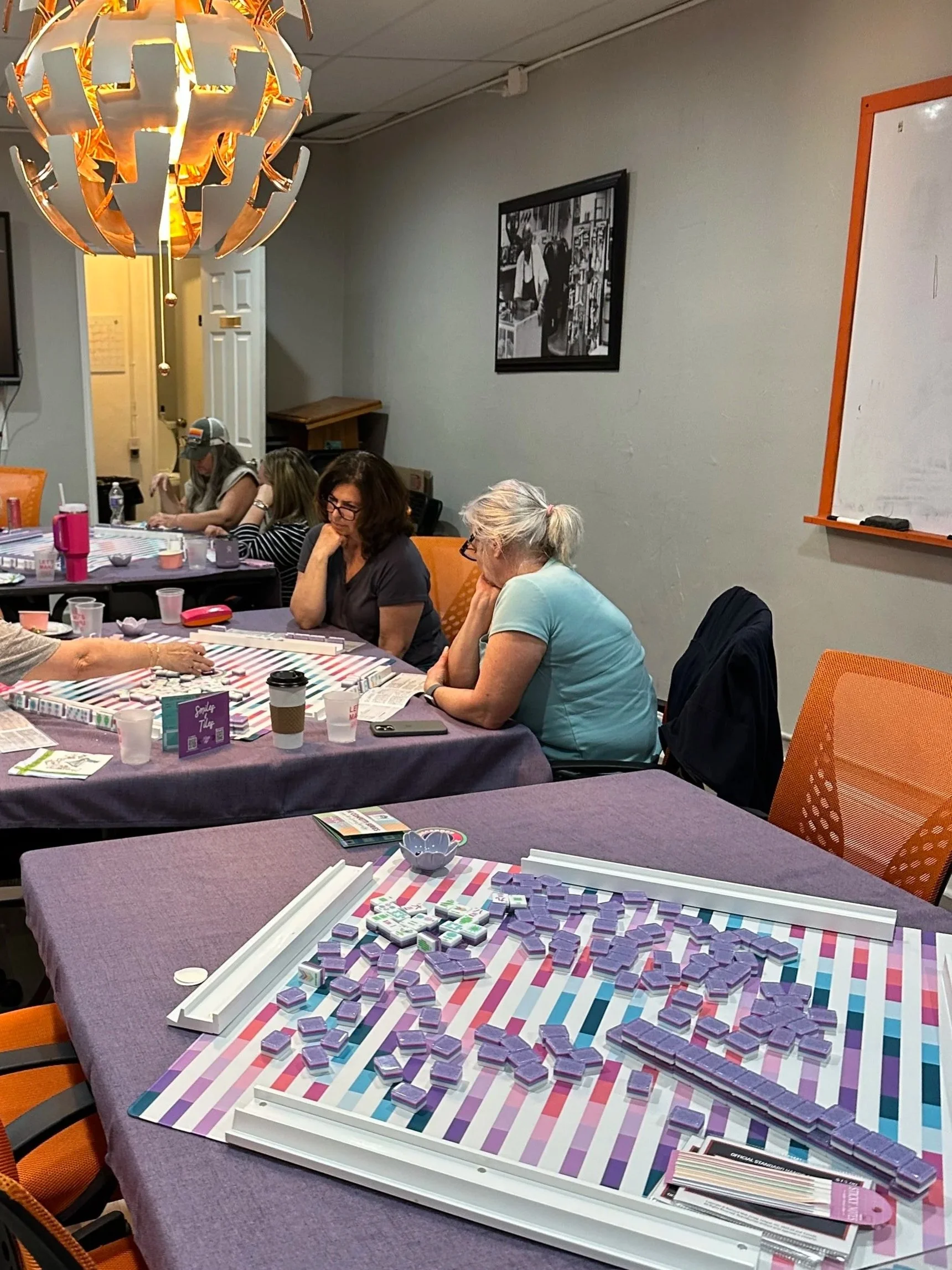 People playing a game of Scrabble at a table in a community room, with some participants focused on the game and others talking, while game pieces and tiles are spread out on the table.