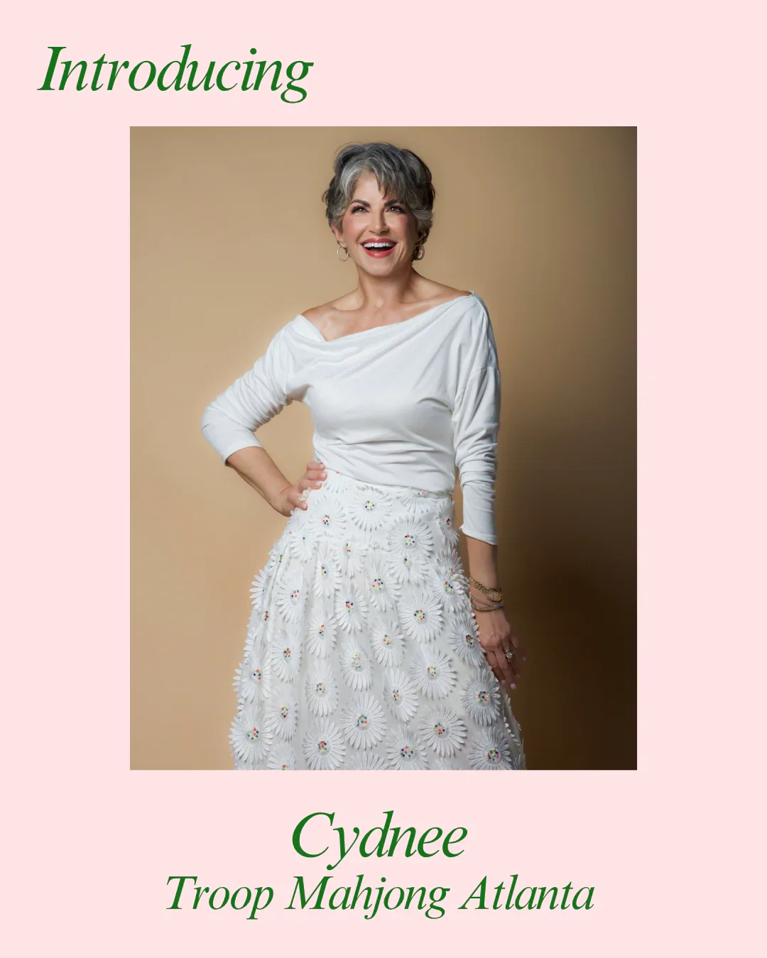 A smiling woman with short gray hair wearing a white top and a white skirt with daisy flower designs, standing against a beige background.
