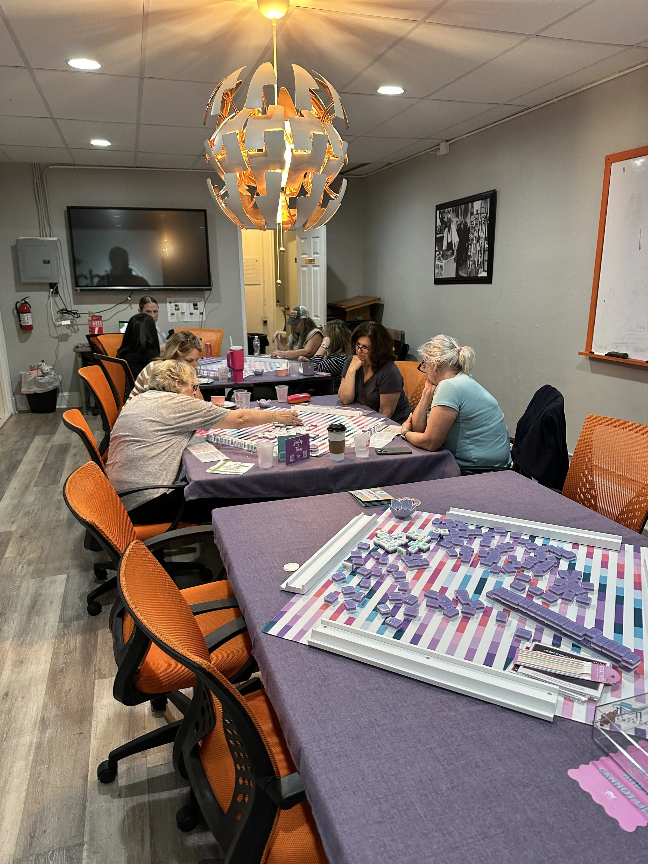 A group of women gathered around a table in a conference room or craft room, working on a project with purple blocks and white frames. The room has orange chairs, a large chandelier, and a whiteboard on the wall.