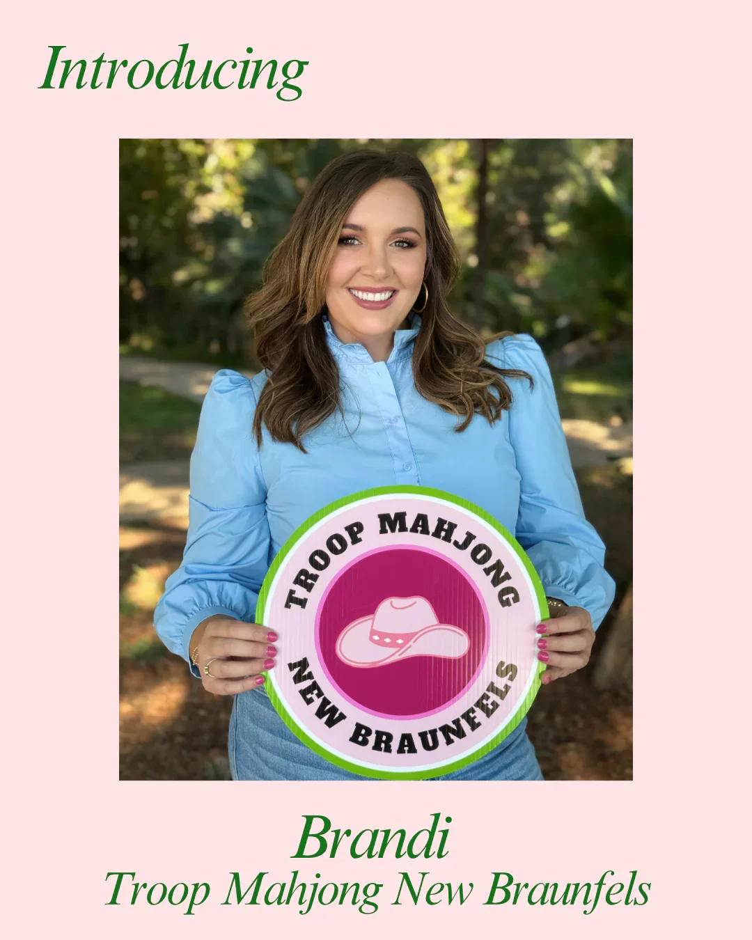 Woman in blue shirt holding a round sign with the text 'Troop Mahjong New Braunfels' and a pink cowboy hat logo, smiling outdoors.