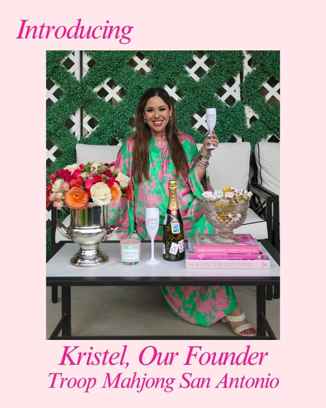 A woman sitting on a white outdoor sofa, holding a champagne flute, surrounded by flowers, champagne, and books, with a green leafy wall behind her. She is smiling and dressed in a colorful pink and green dress.