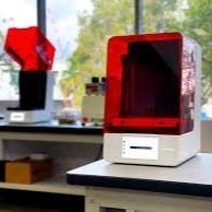 A red and white 3D printer on a white table with a computer monitor in the background.
