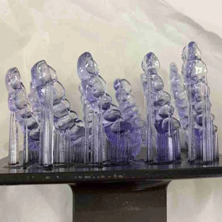 Close-up of a glass sculpture with multiple tall, slender, purple-tinted glass pieces of varying heights on a reflective black base.