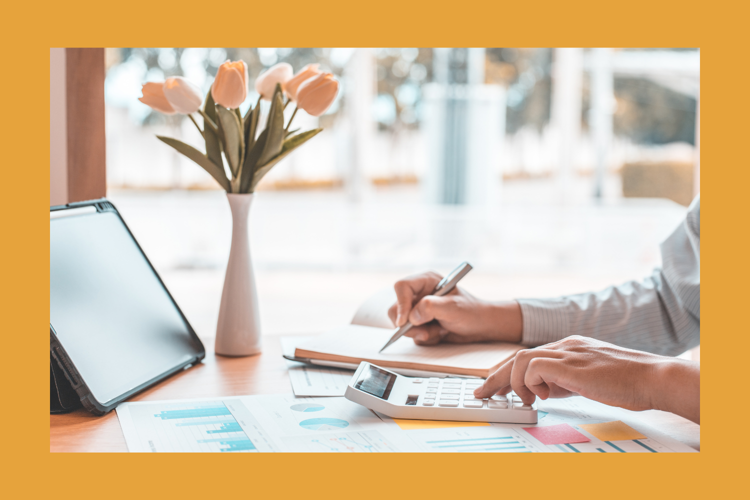 Person using calculator and writing in notebook on a desk with a tablet, documents, and a vase with tulips, near a window with blurry outdoor view.