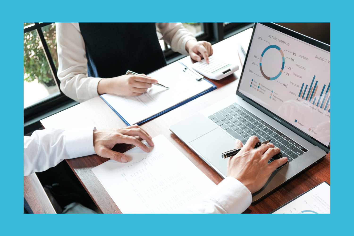 Two people working at a desk with financial documents, a calculator, and a laptop displaying financial charts, in front of a window.