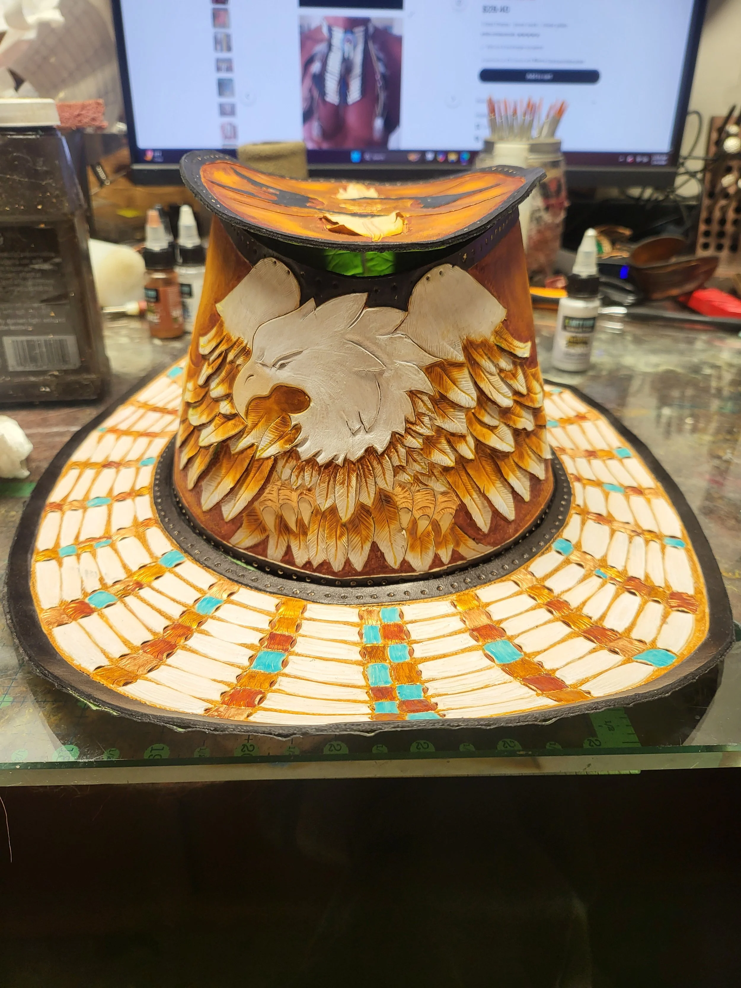 A detailed, colorful handcrafted hat with an eagle design, on a worktable with art supplies and a computer monitor in the background.