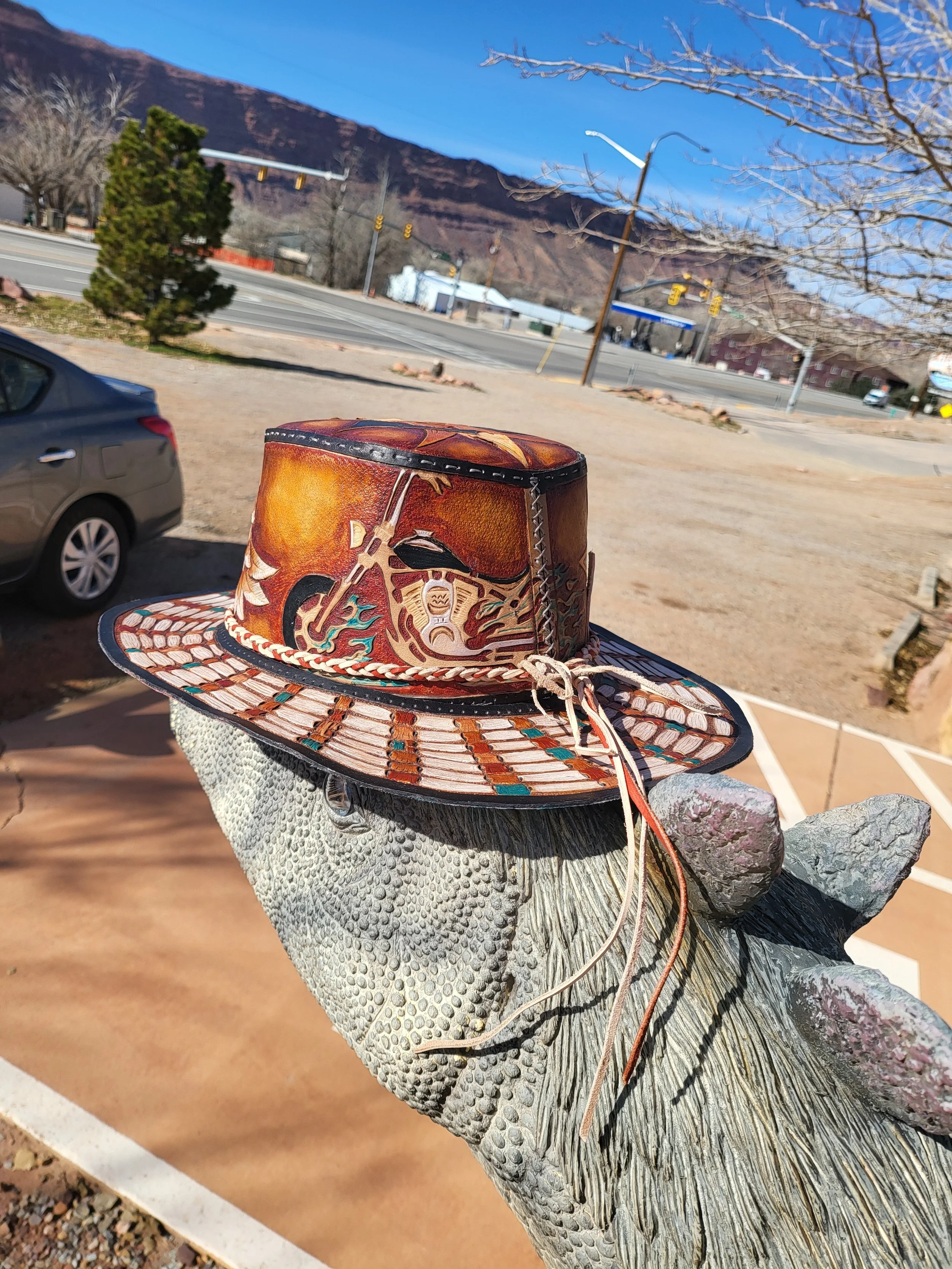 A decorative cowboy hat with painted cowboy and mountain designs, placed on a desert animal sculpture outside.