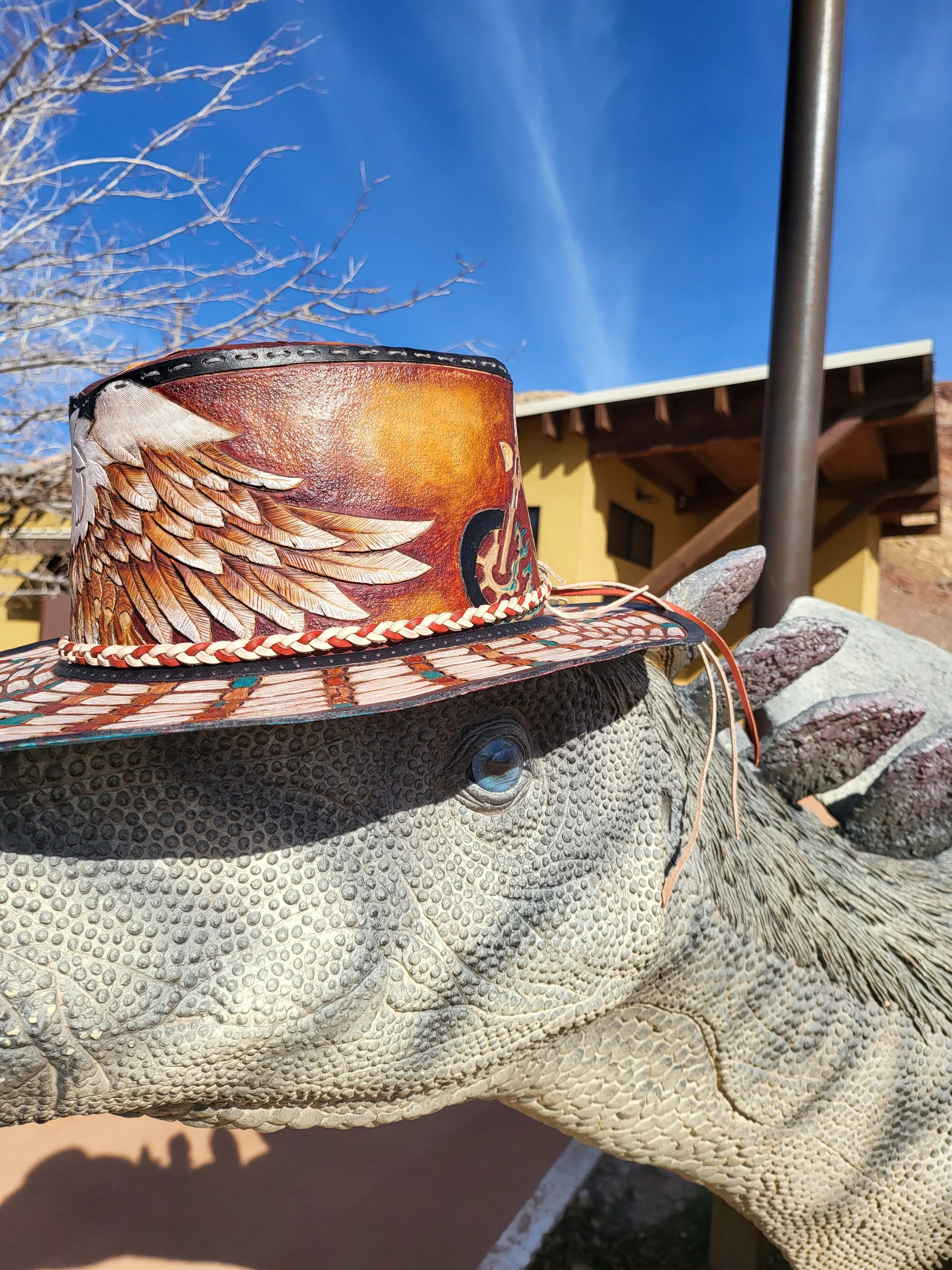 A rhinoceros sculpture with a decorated cowboy hat on its head, set outdoors with a building, a pole, a tree, and a clear blue sky in the background.