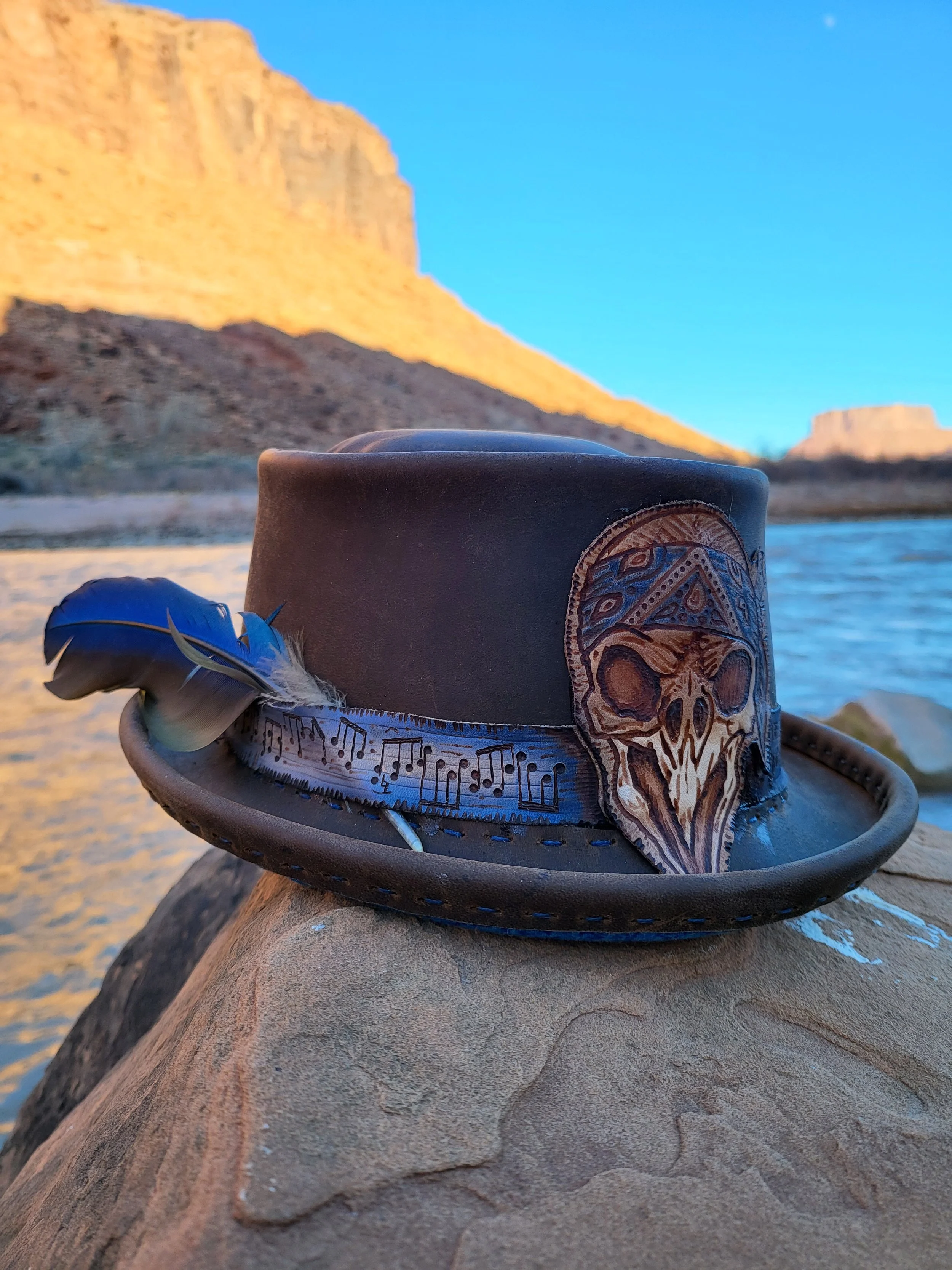 A leather hat with a decorative skull patch and a feather, resting on a rock by a river with cliffs in the background at sunset.