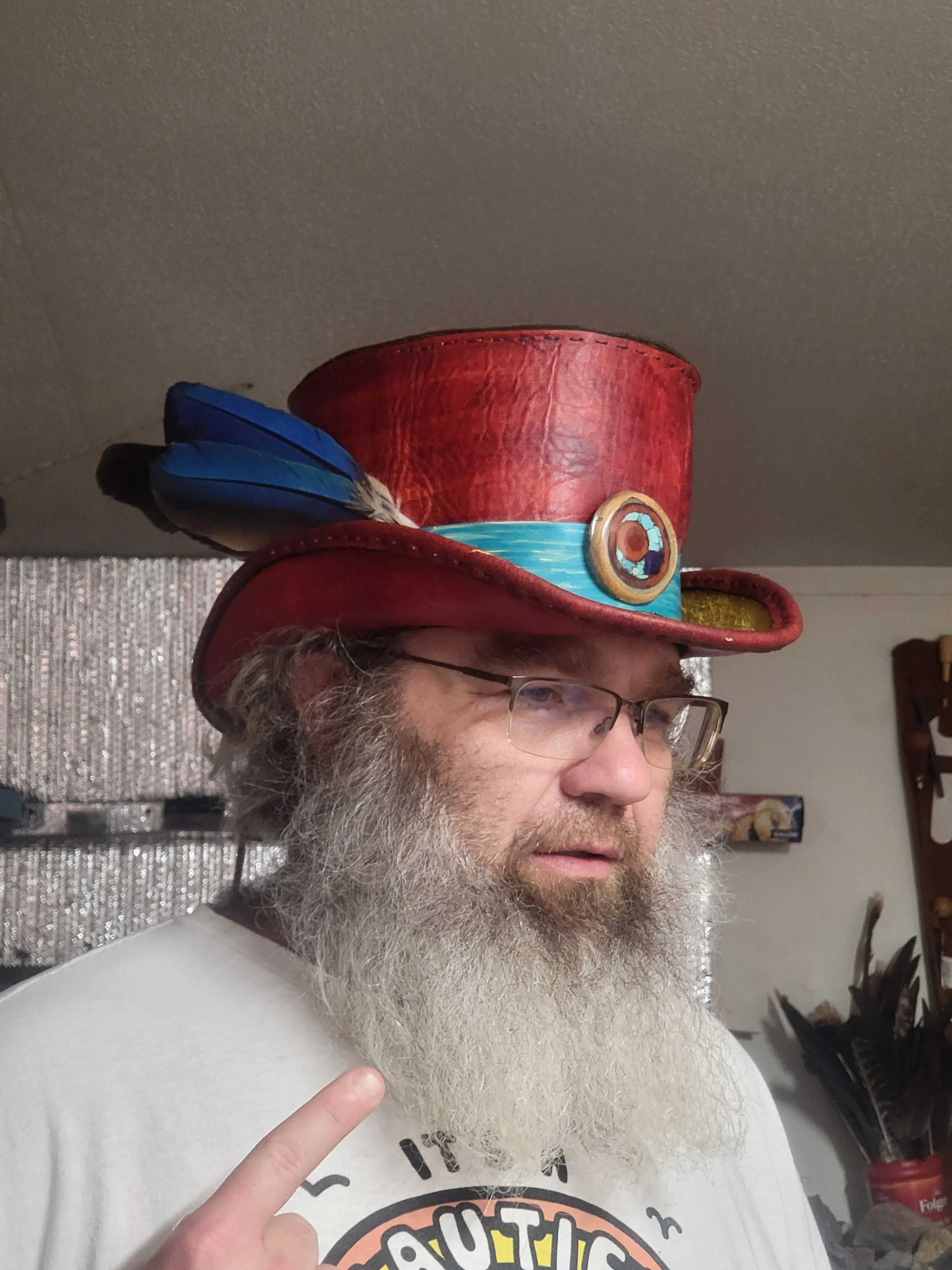 A man wearing glasses and a white t-shirt with a graphic design, pointing and wearing a tall, red top hat decorated with blue feathers, a blue band, and a circular emblem.