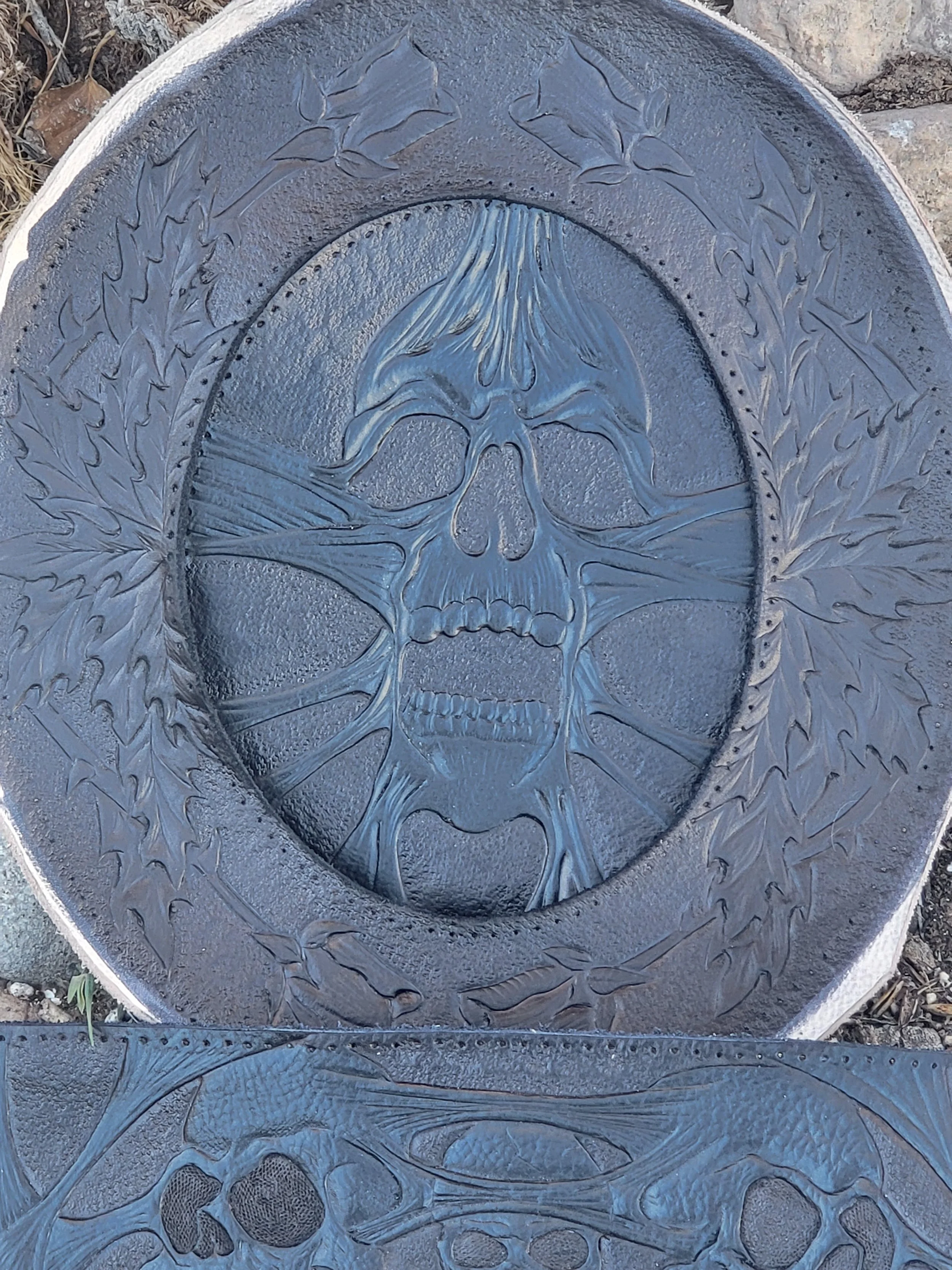 Decorative metal plate with a skull design surrounded by leaf patterns.