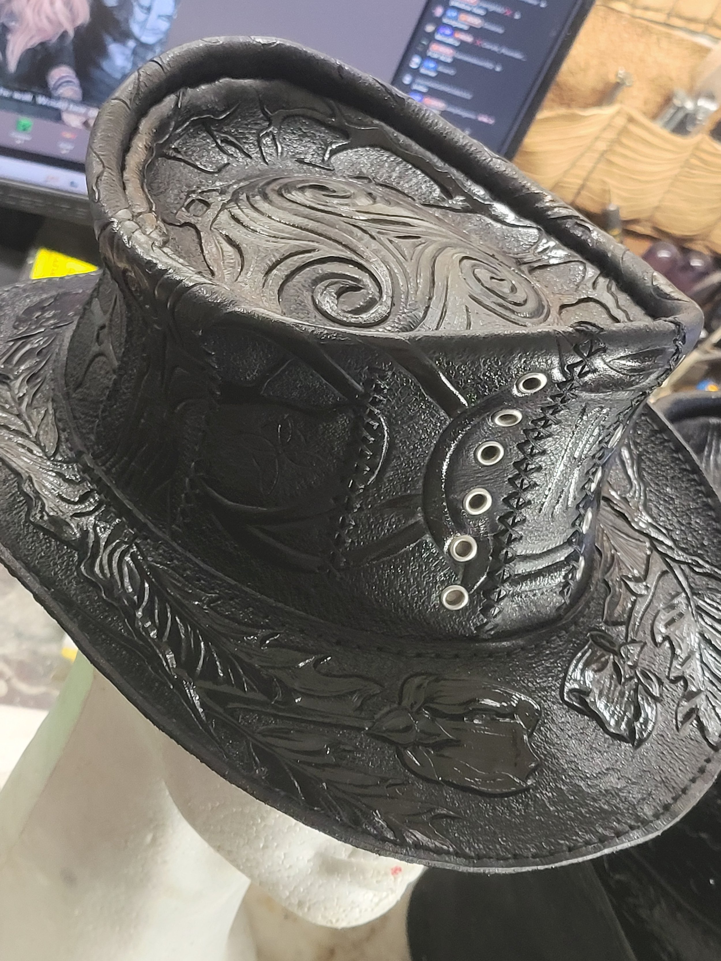 Close-up of a black leather cowboy boot with intricate tooled designs, including a spurred heel, eyelets, and embossed floral patterns, with a computer screen and shelves in the background.