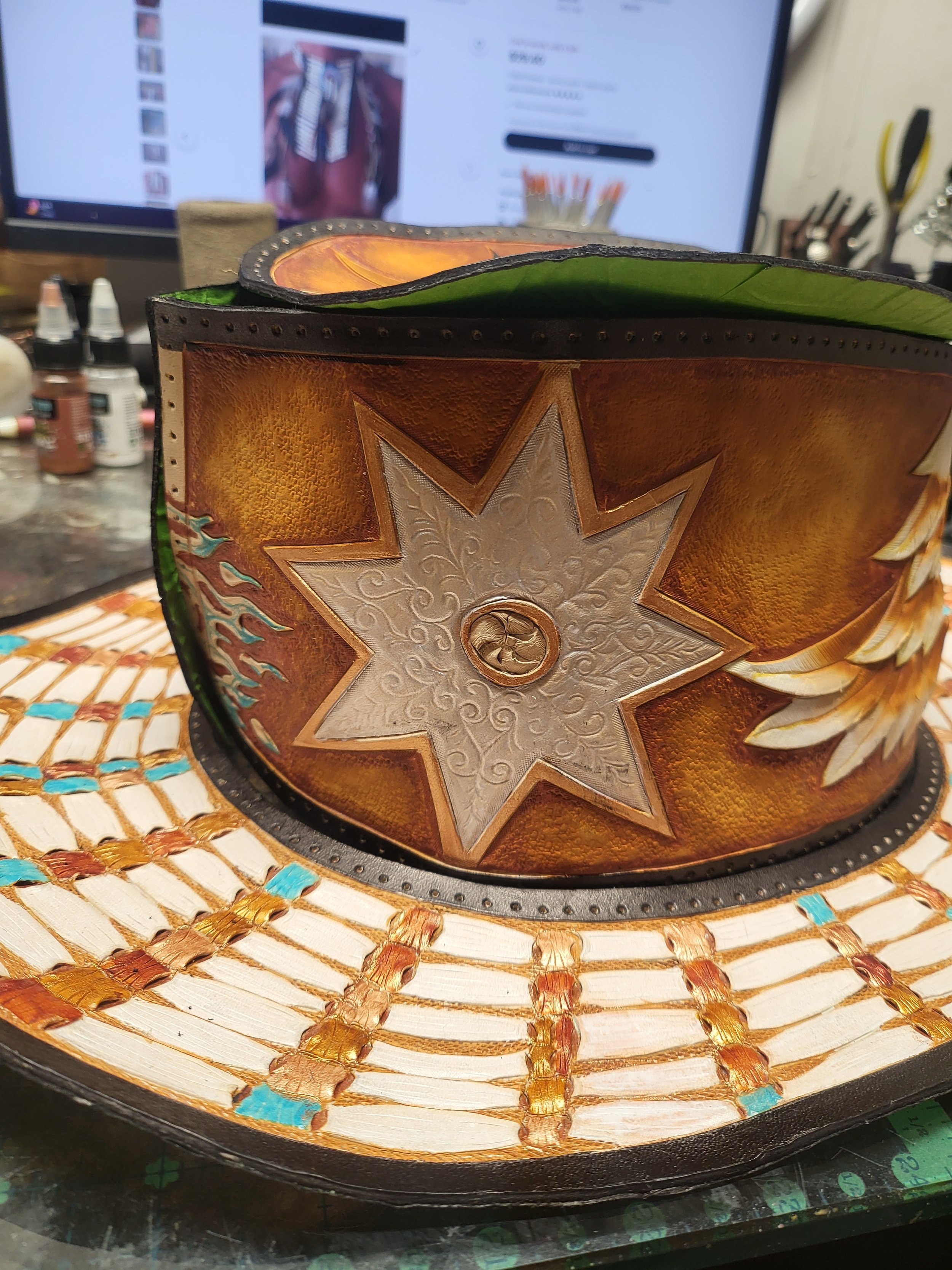 A decorated cowboy hat with a leather band featuring a star emblem and painted designs, placed on a worktable with painting supplies in the background.