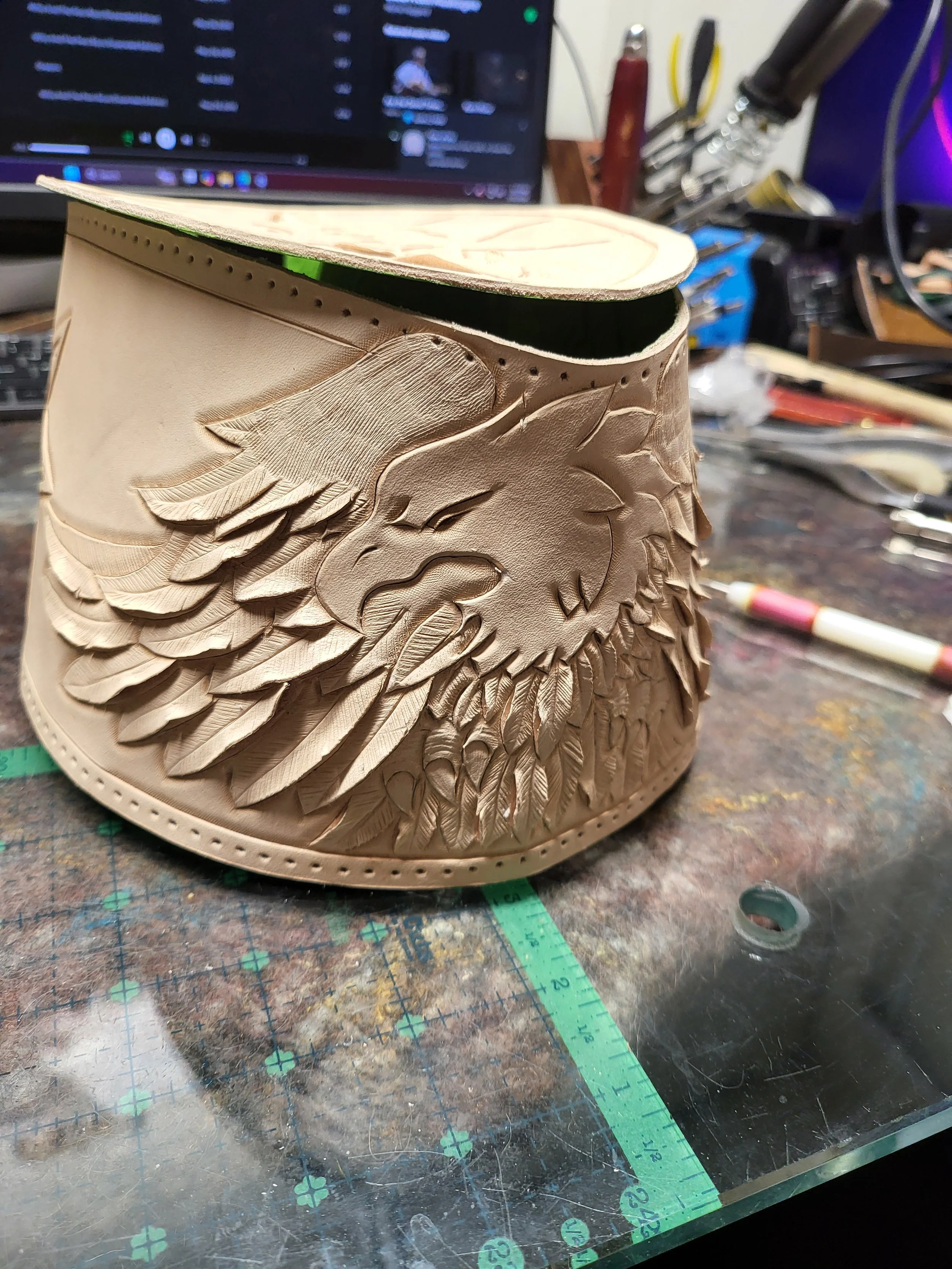 A work-in-progress leather shoe with a detailed eagle head carving, situated on a cluttered workbench with tools and a computer monitor in the background.