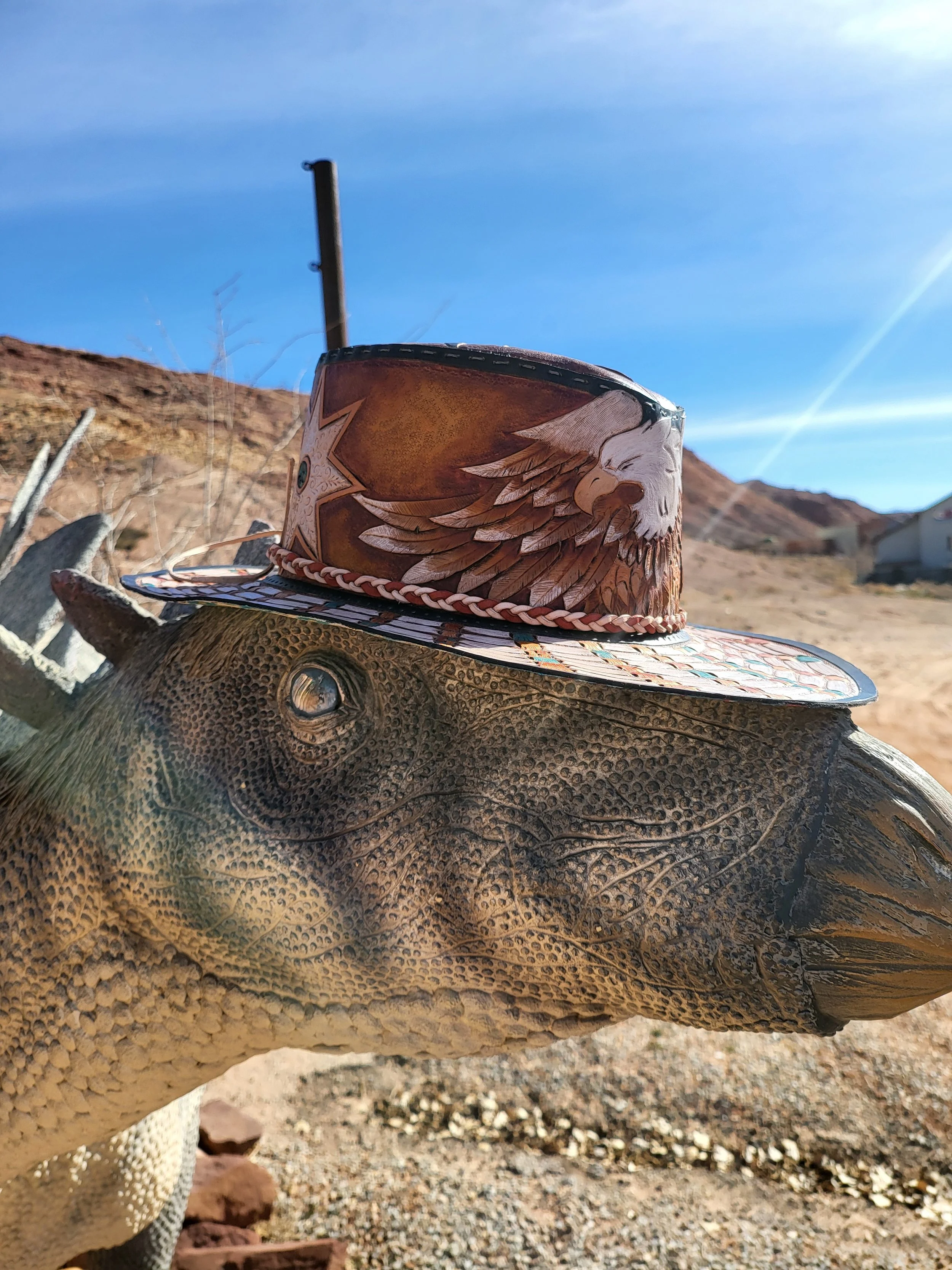A large lizard with scaled skin wearing a decorative cowboy hat with a painted eagle and star design, set against a desert landscape with mountains and a clear blue sky.