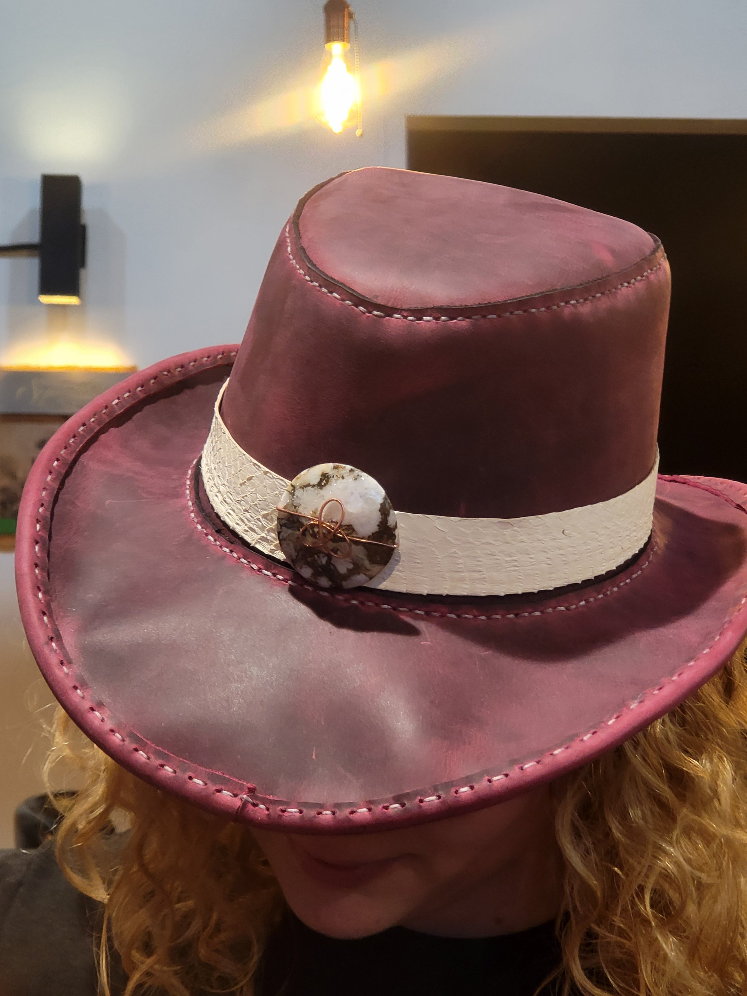 A person wearing a large pink cowboy hat with a white band and decorative stone pin, with curly blonde hair visible underneath, in a dimly lit room.