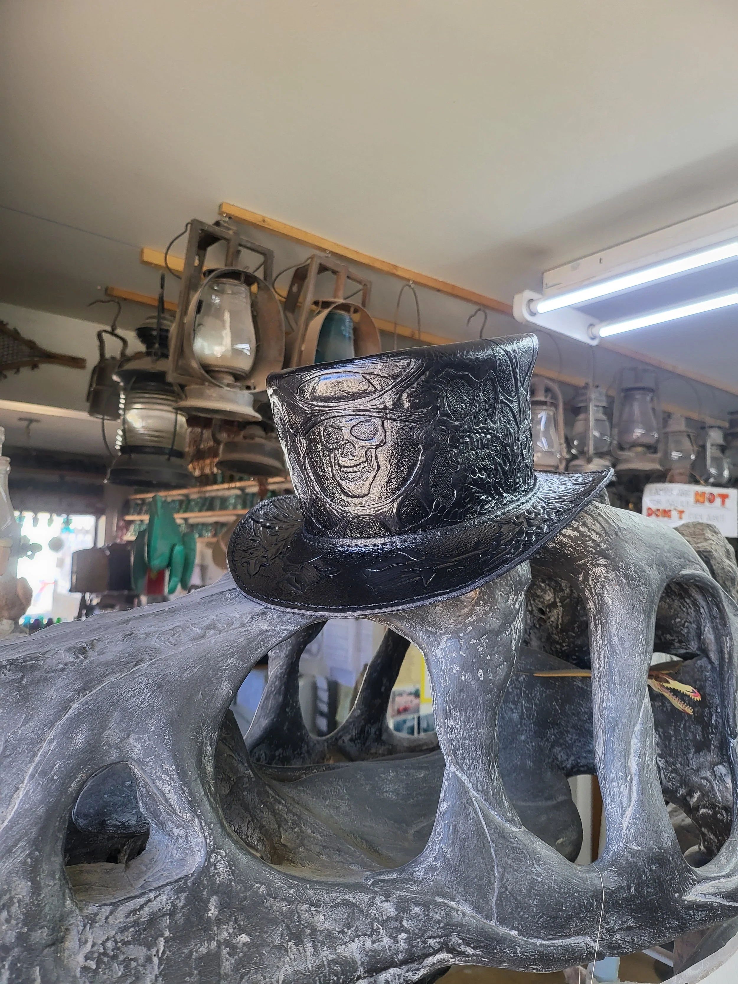 A decorative black top hat with a skull and snake design, placed on a weathered, hollow, metallic sculpture with multiple openings, inside a store with hanging lanterns and signage.