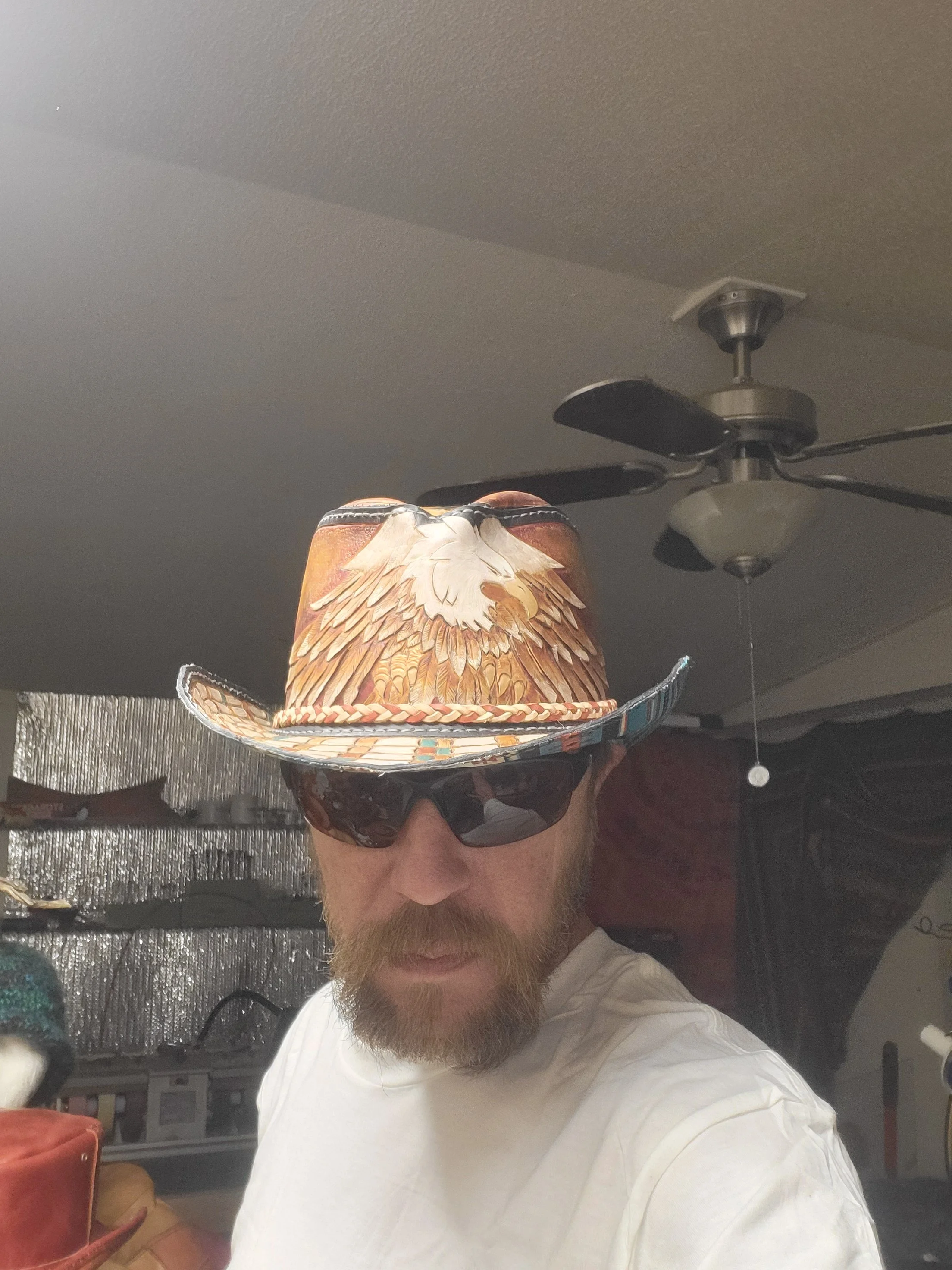 Man with a beard wearing sunglasses and a cowboy hat with an eagle design, indoors with a ceiling fan overhead.
