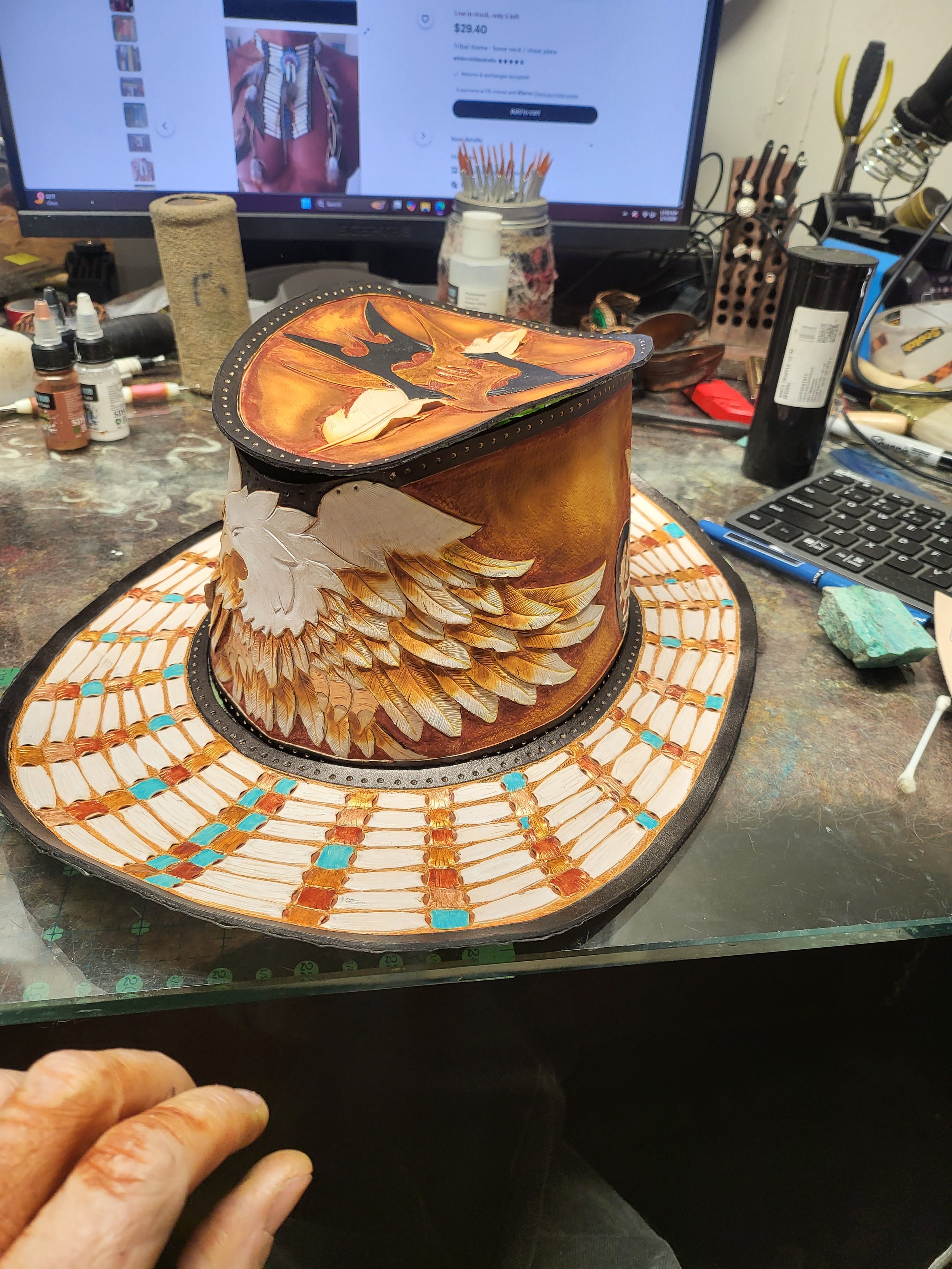 Painted cowboy hat with eagle feather design on the brim and a sunset scene on the top, on a cluttered work table.