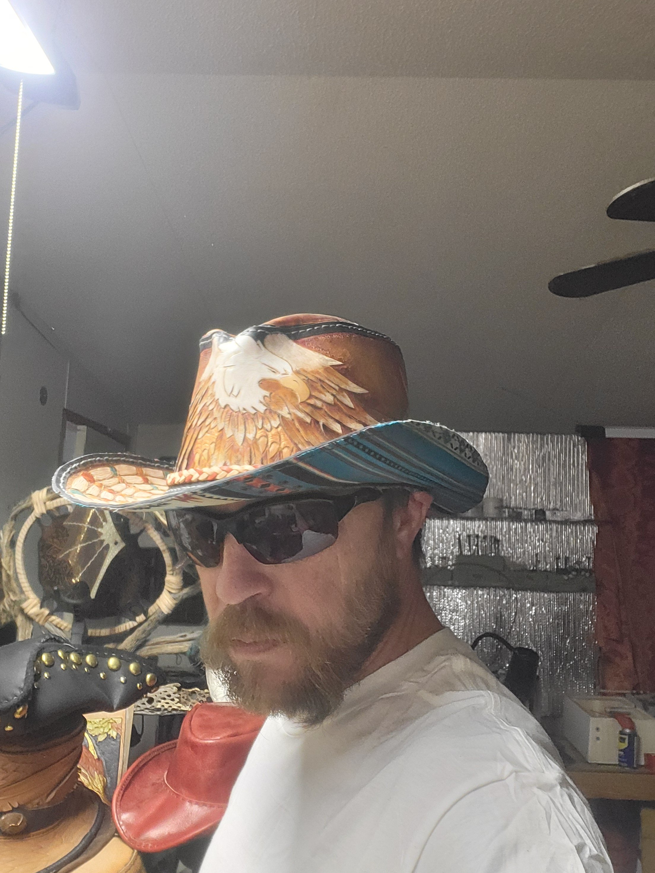 A man wearing dark sunglasses and a cowboy hat with an eagle design, taking a selfie in an indoor setting.