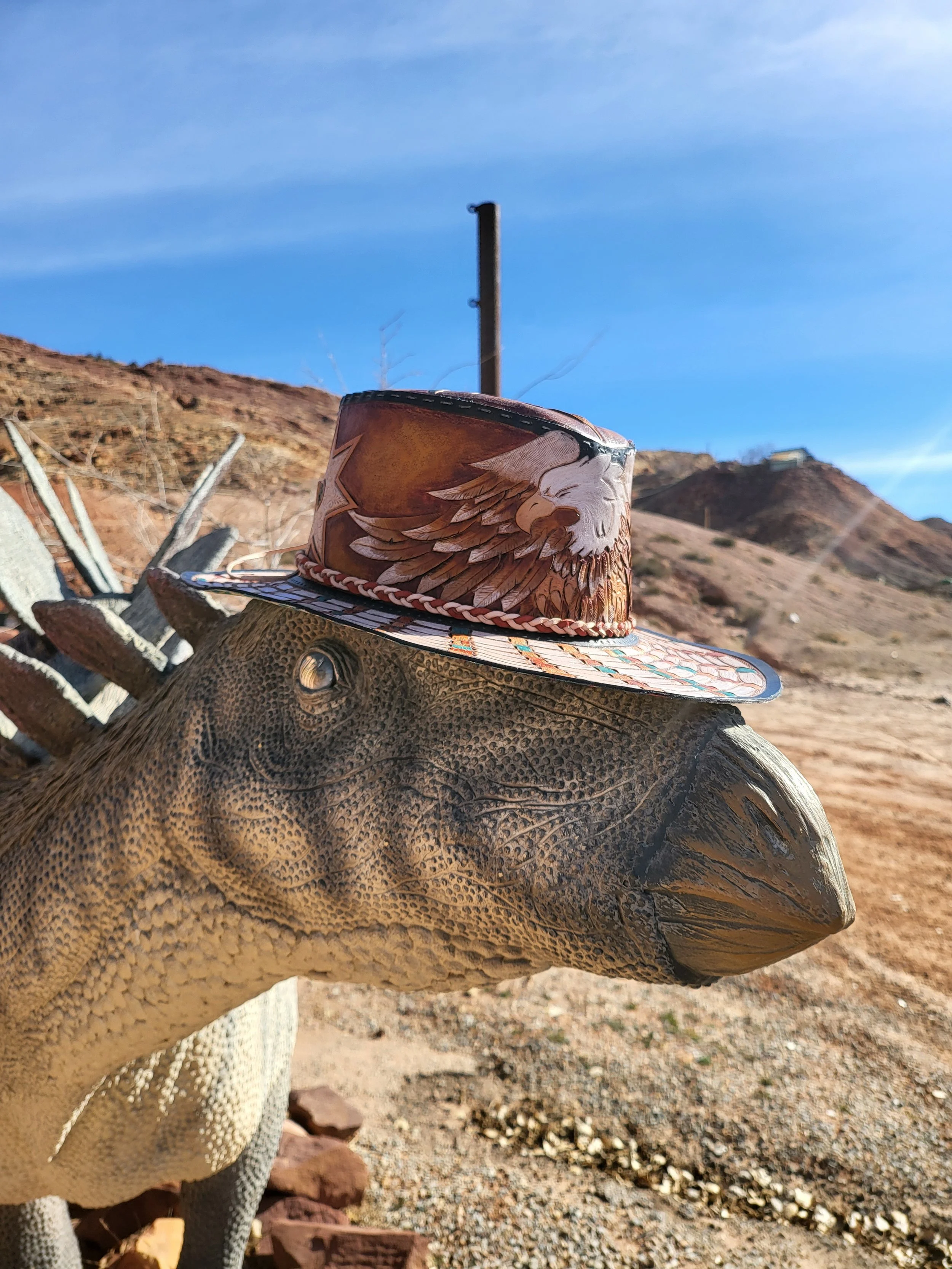 A dinosaur sculpture wearing a cowboy hat with an eagle design, set outdoors with desert hills and a clear blue sky in the background.