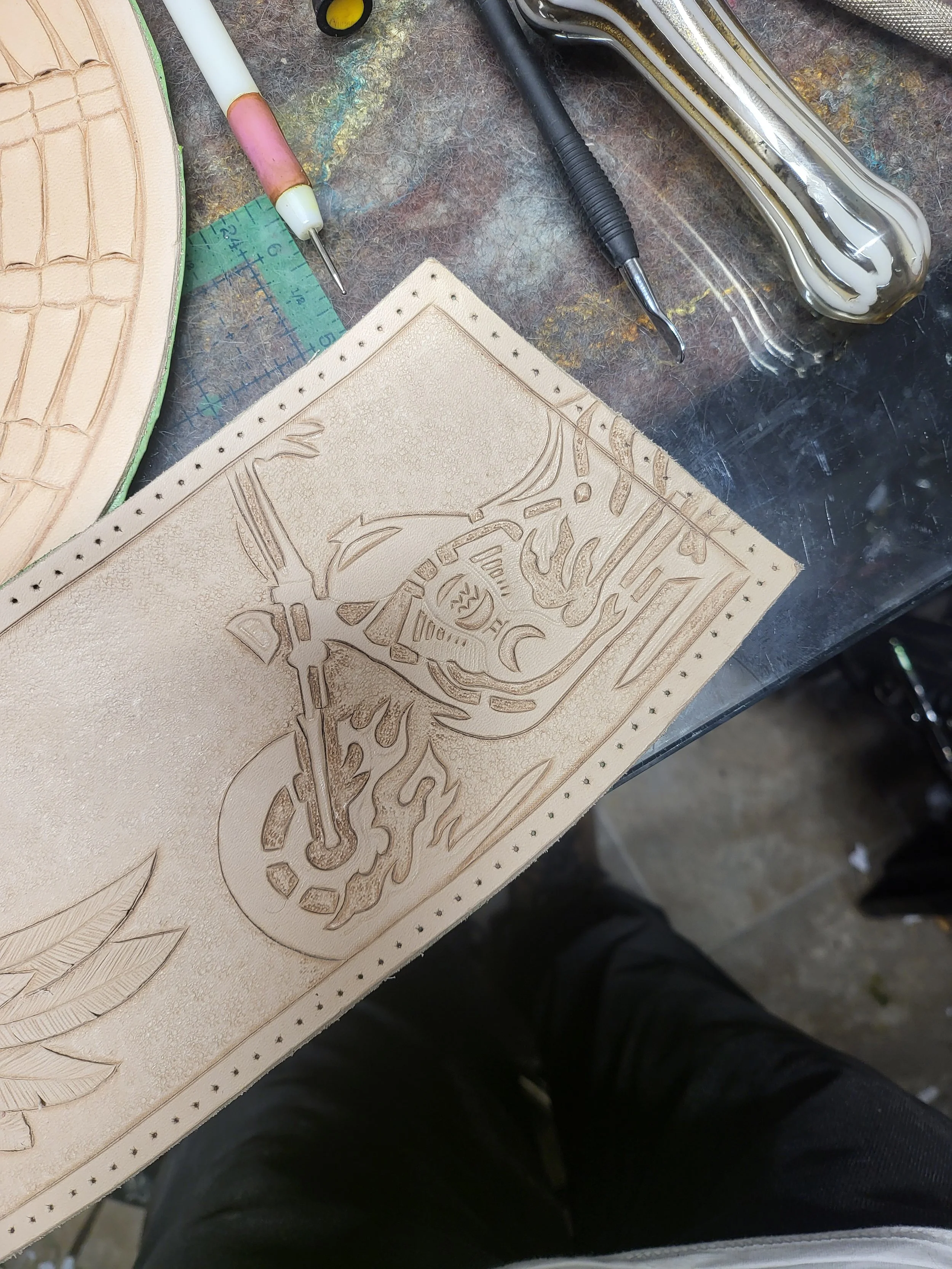 Leatherworking tools and a piece of leather with a carved design of a sword, flames, and Arabic script on a workbench.