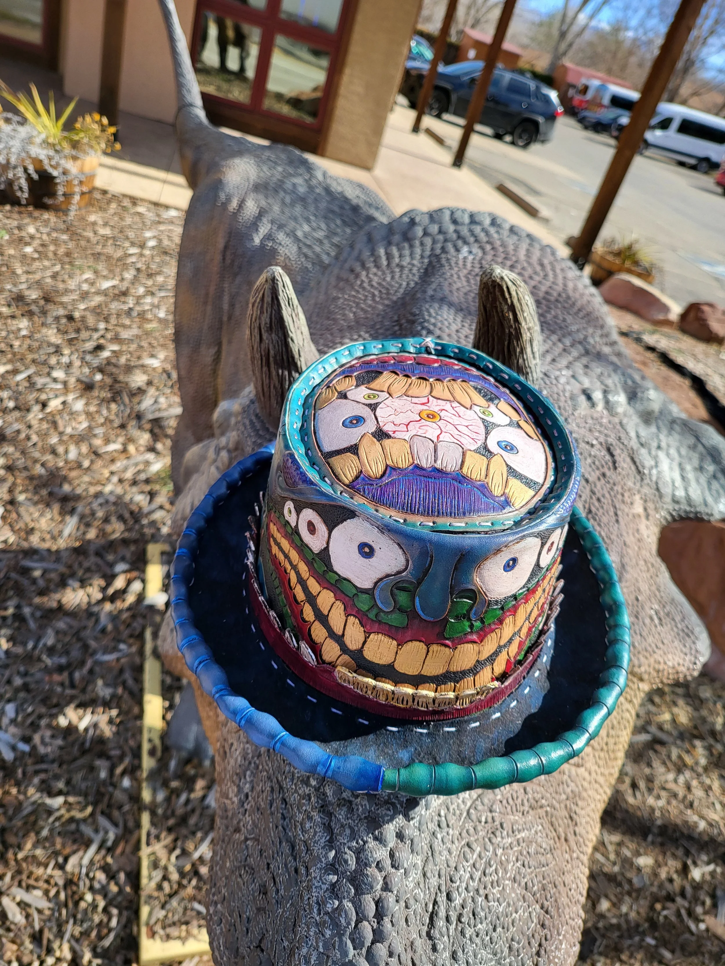 Painted face mask with multiple eyes and teeth, placed on a decorative headband, resting on the back of a large lizard sculpture outdoors.