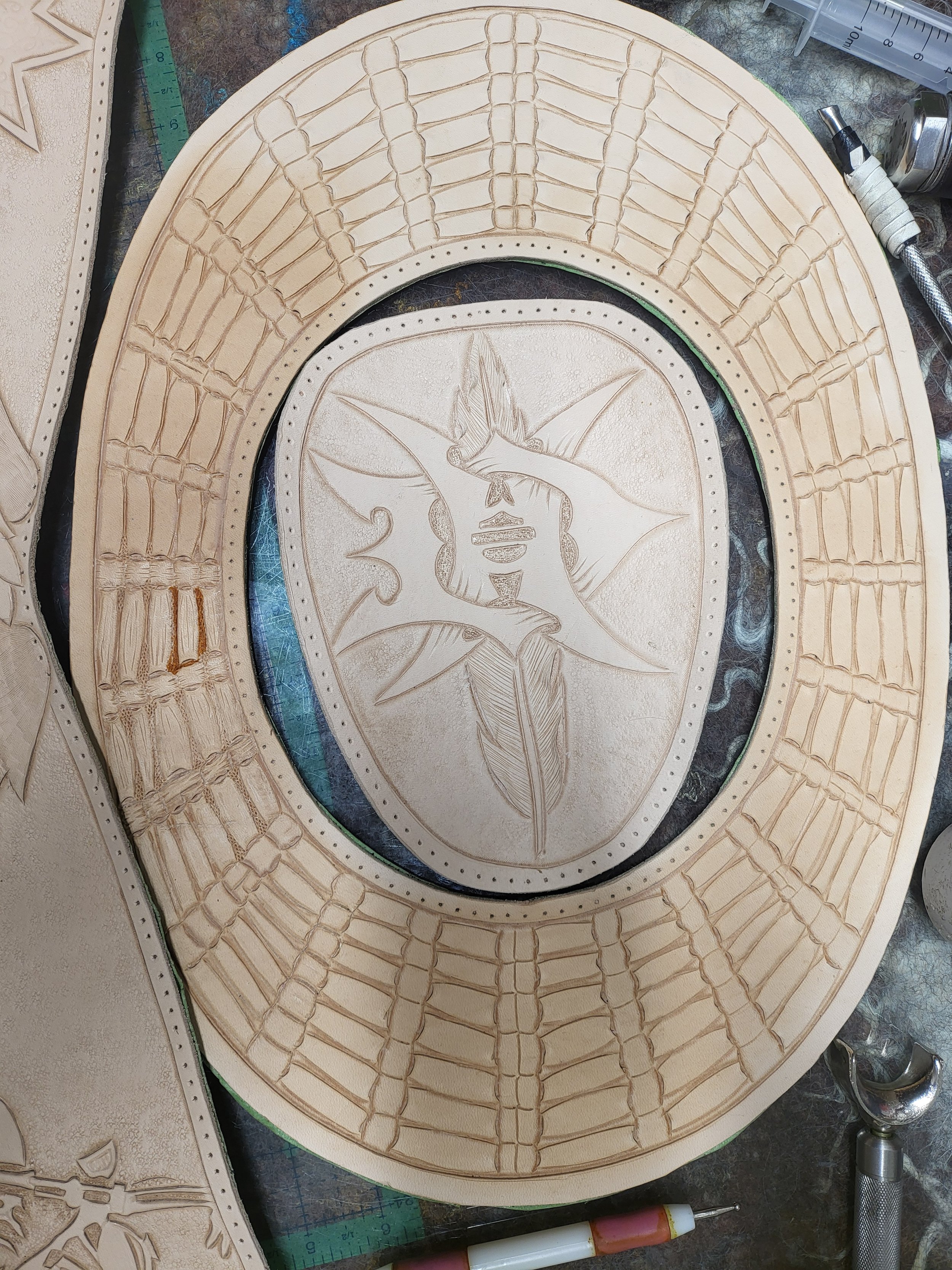 A carved leather piece featuring a design of a hummingbird and a flower, with a straw flower at the bottom. The leather is beige with detailed embossing, surrounded by a decorative border.