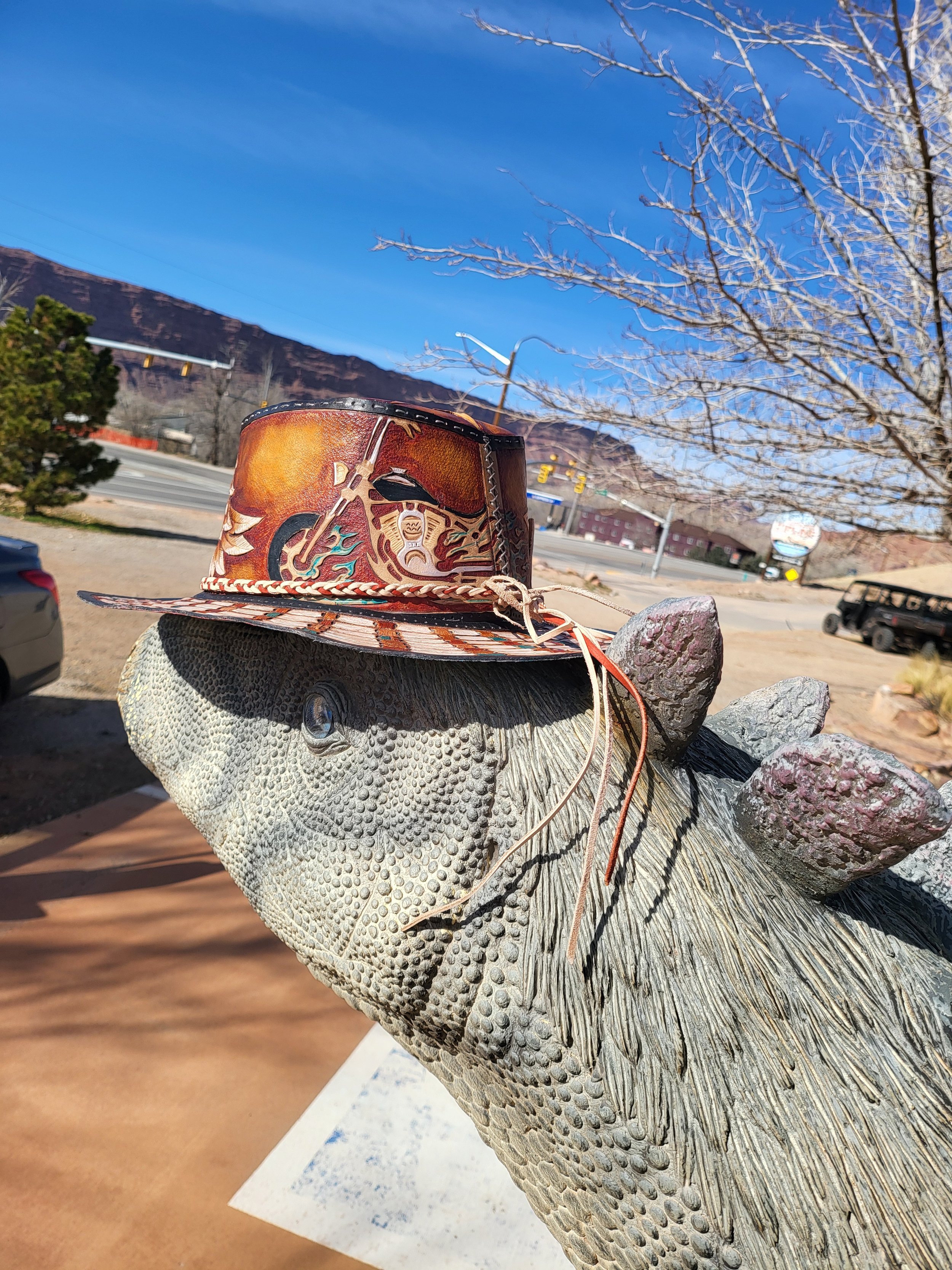 A decorative pig sculpture with a cowboy hat placed on its head, outdoors with a mountain, trees, and a blue sky in the background.