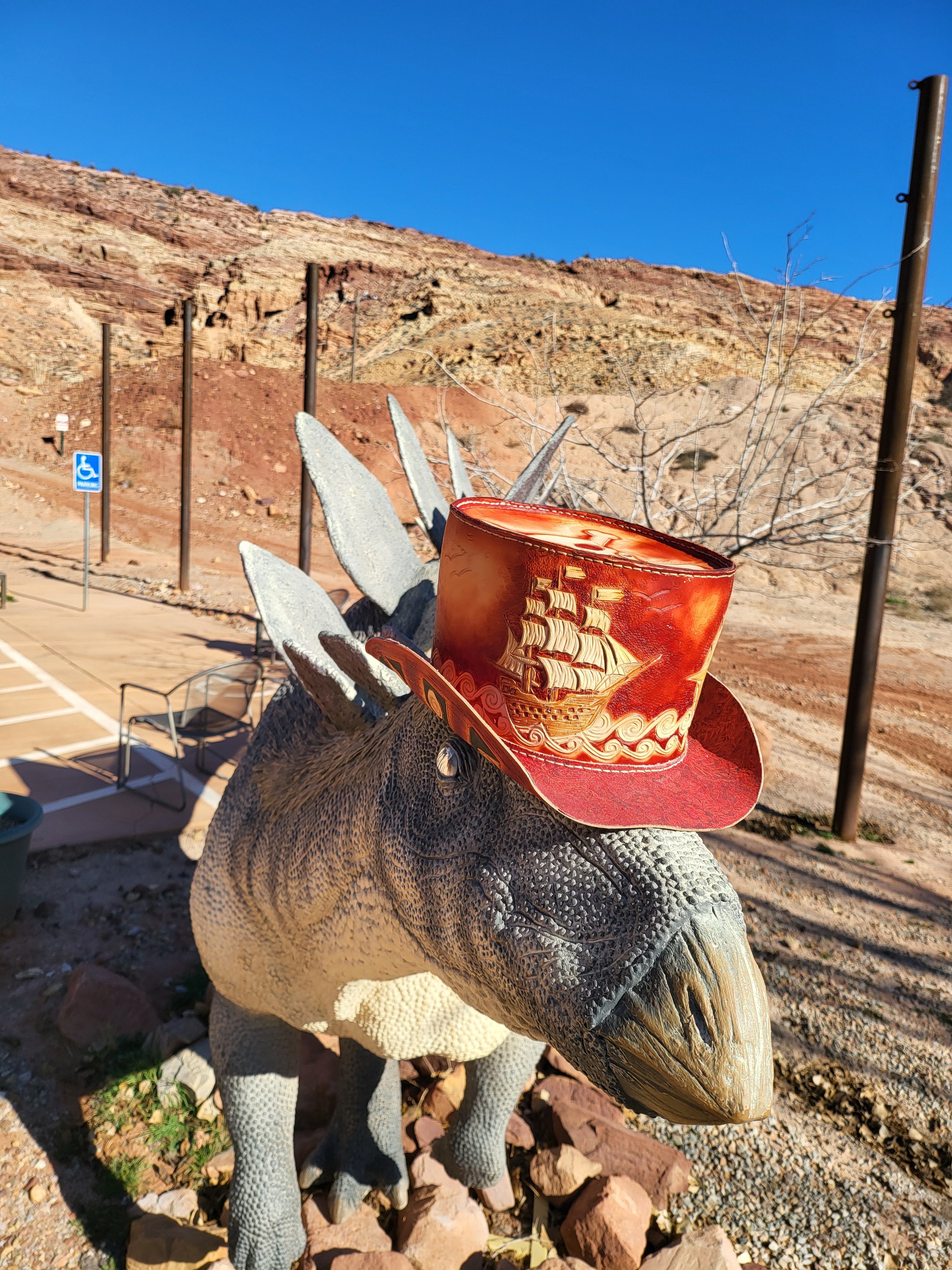 A dinosaur statue wearing a red cowboy hat adorned with a sailing ship design, situated outdoors against a desert landscape with rocky hills, a clear blue sky, and some parking areas.