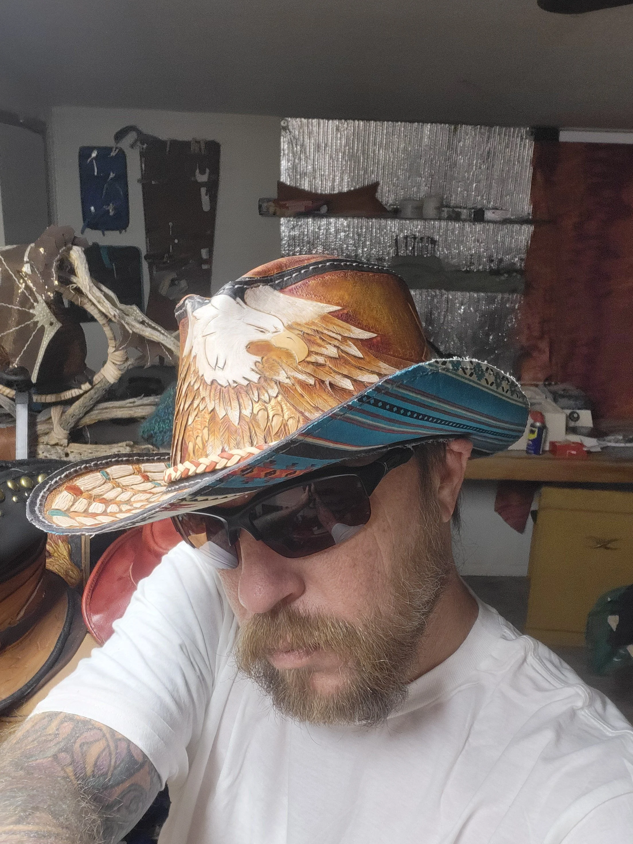 A man wearing a colorful cowboy hat with an eagle design, sunglasses, and a white T-shirt. He has a beard and tattoos on his arm, and the background includes various objects and equipment, suggesting a workshop or studio environment.