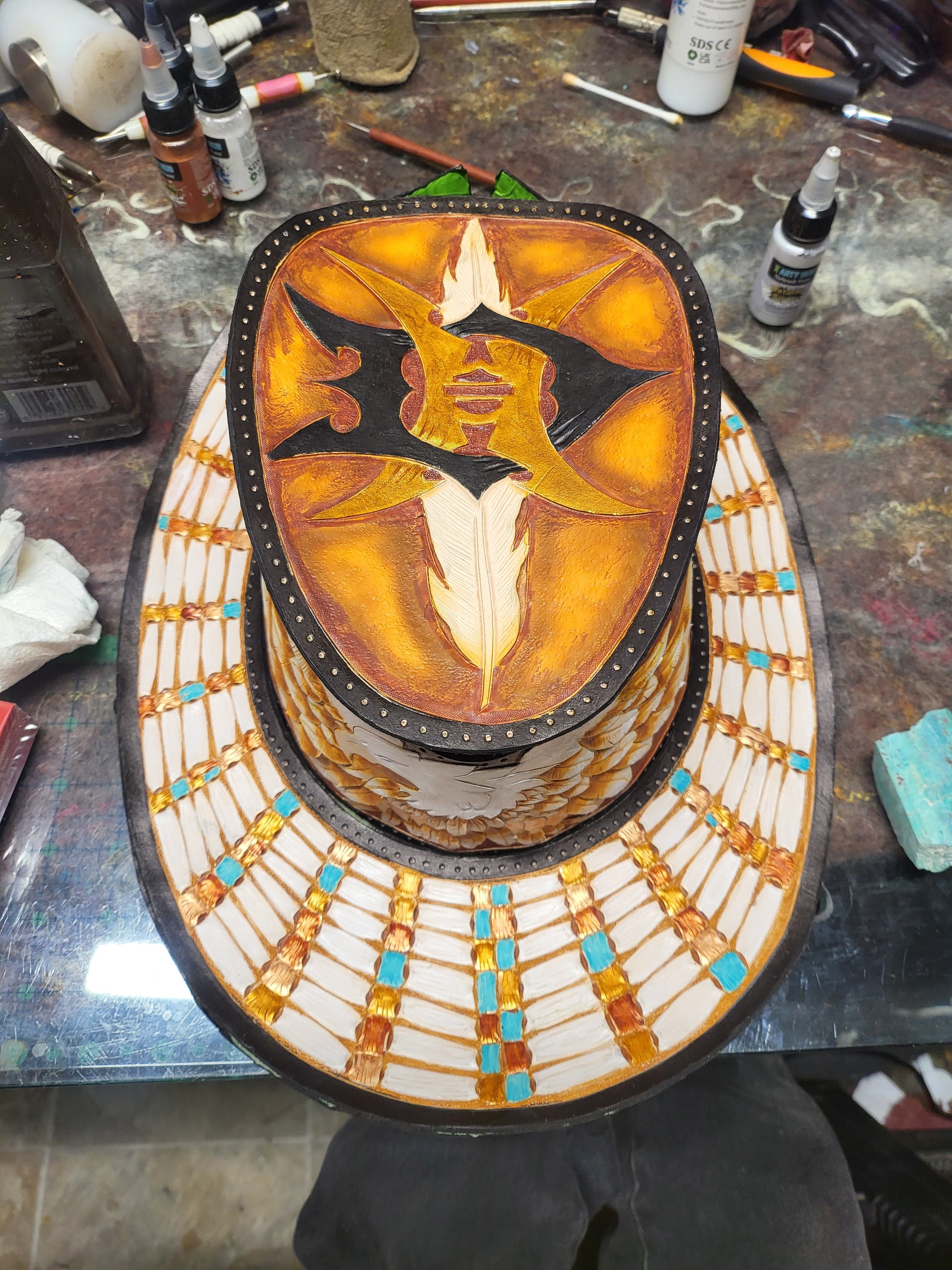 Hand-painted decorative hat featuring intricate designs with warm colors, black accents, and a feather motif, placed on a cluttered workbench with paints and tools.