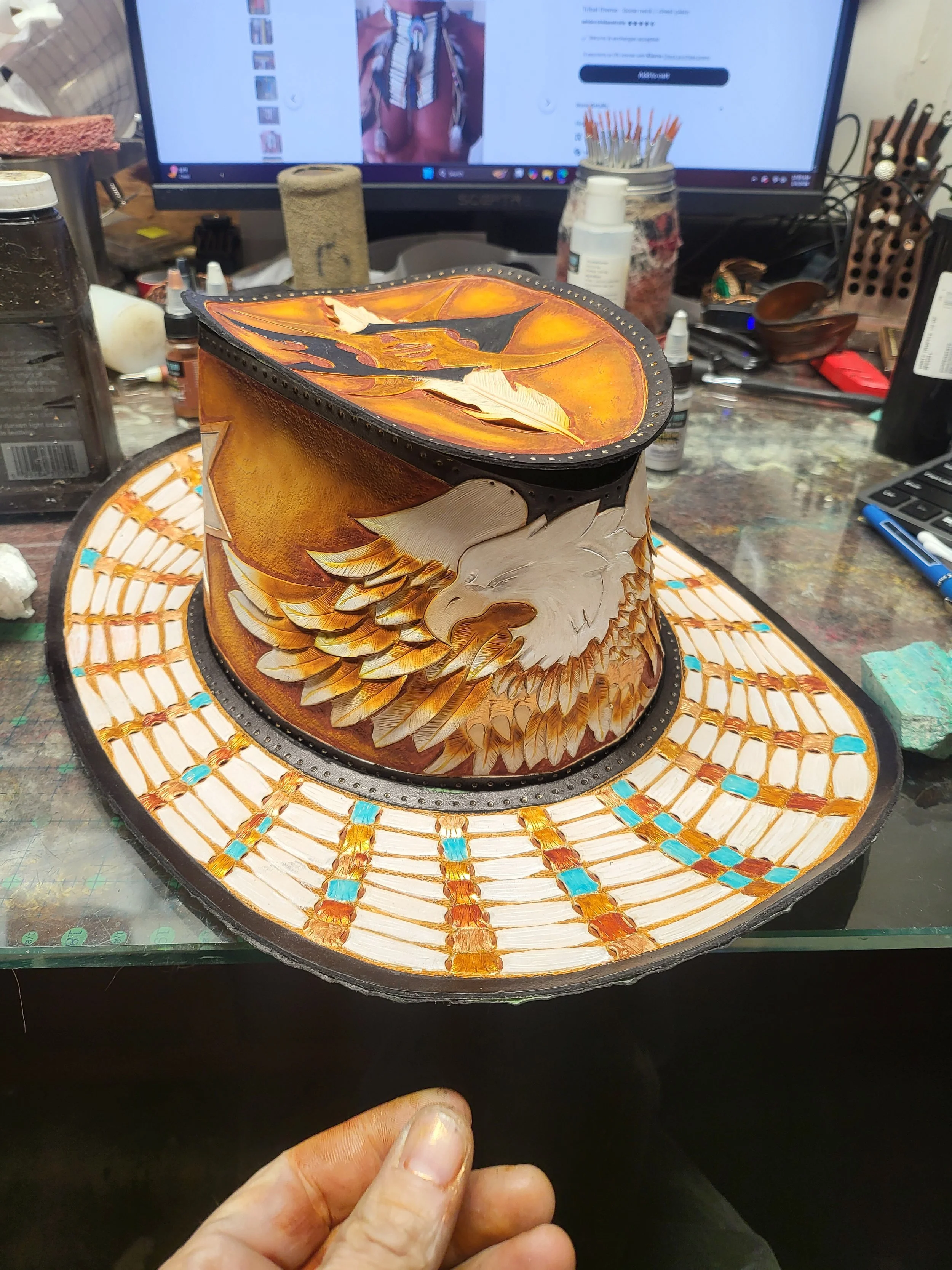 Handcrafted leather hat with an eagle design, featuring detailed feather illustrations and a decorative brim with geometric patterns in orange, white, and blue colors, on a worktable with art supplies and a computer monitor in the background.