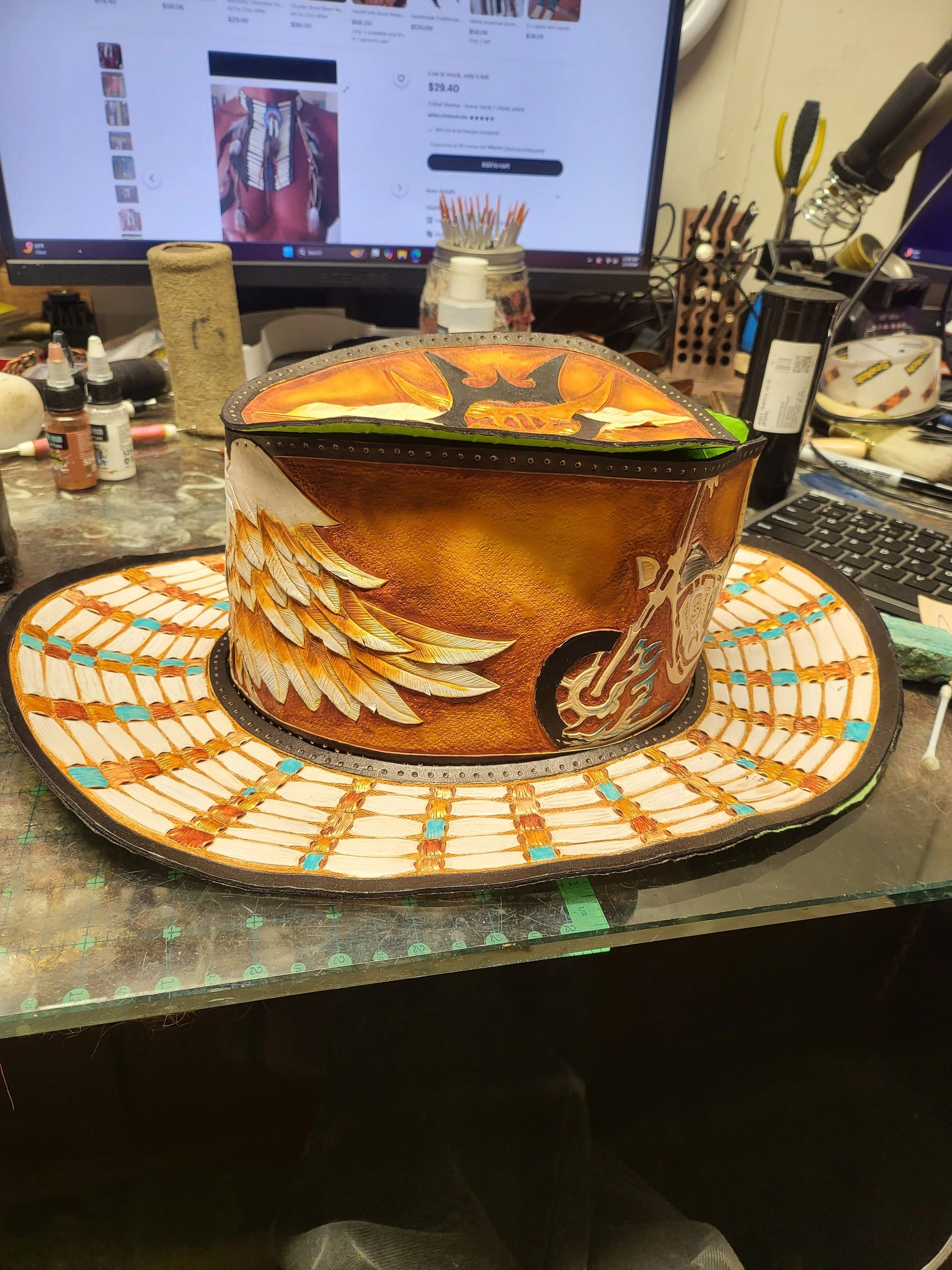 A handcrafted leather cowboy hat with painted artwork featuring an eagle's wings, a guitar, and a skull, placed on a work desk in front of a computer monitor.