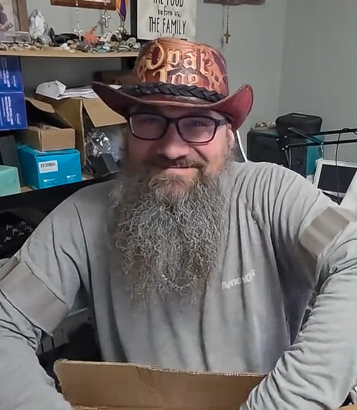 A man with a long beard wearing glasses and a colorful cowboy hat, sitting at a cluttered desk in a room.